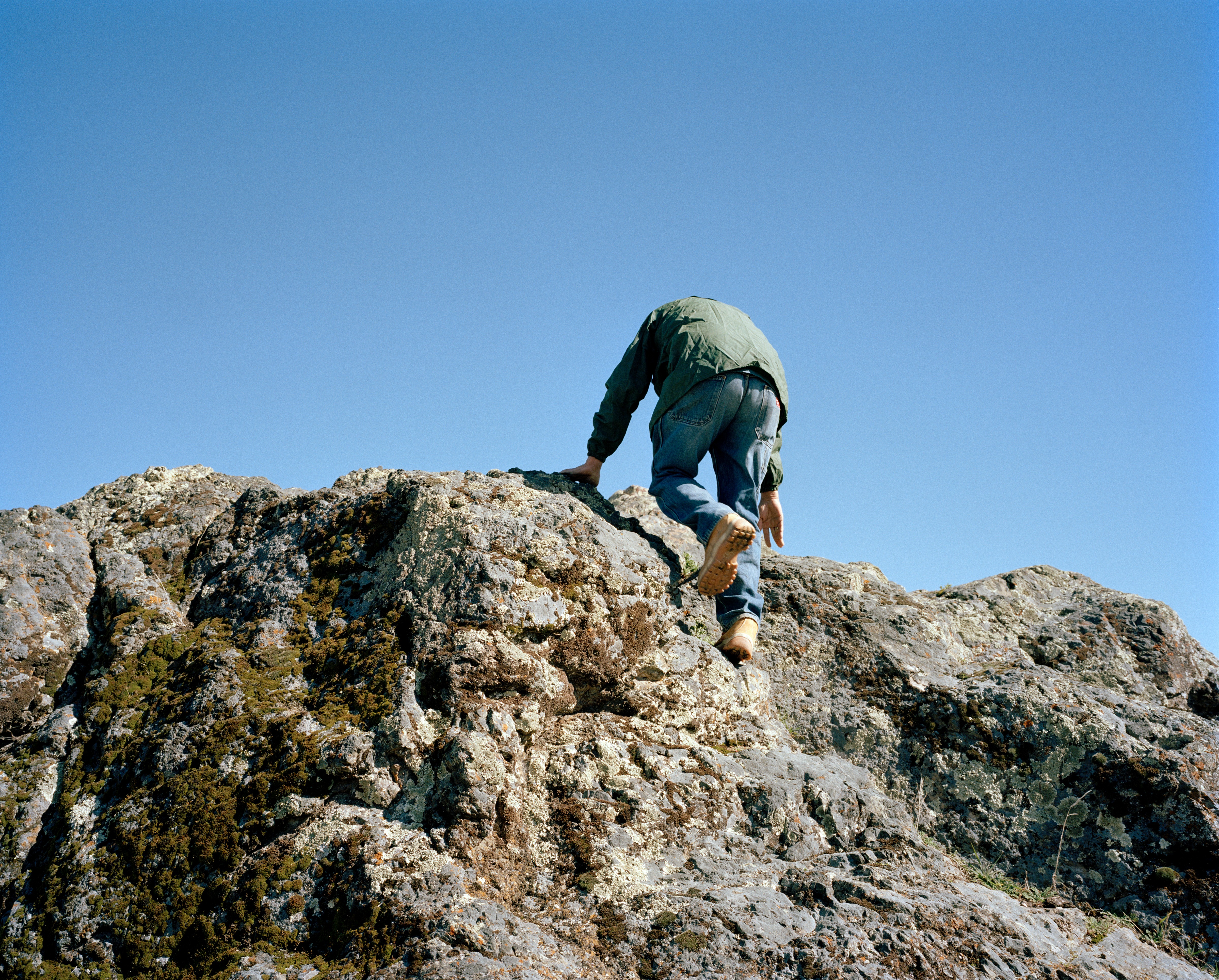 A photograph of a man climbing a rocky hill. The viewpoint is from behind and the sky is clear and blue.
