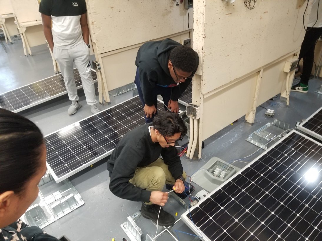 High school students crouch toward the ground in order to plug cords into hardware for solar panels, which surround the students in a workshop.