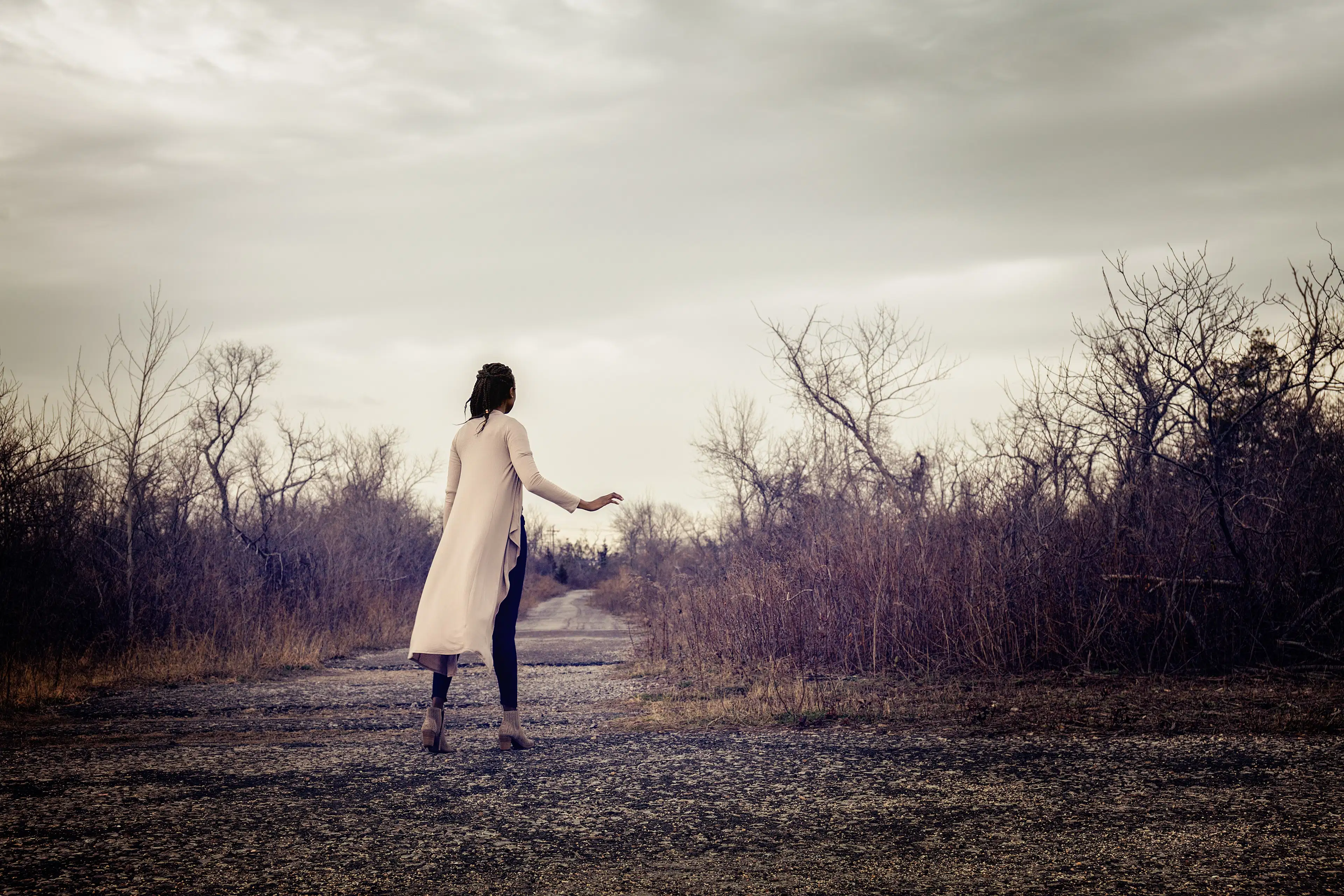 A black woman stands with her back to us. She is wearing a long sweater and hands are up in front of her. She is on an old road, surrounded by low brush and trees, and a cloudy sky.