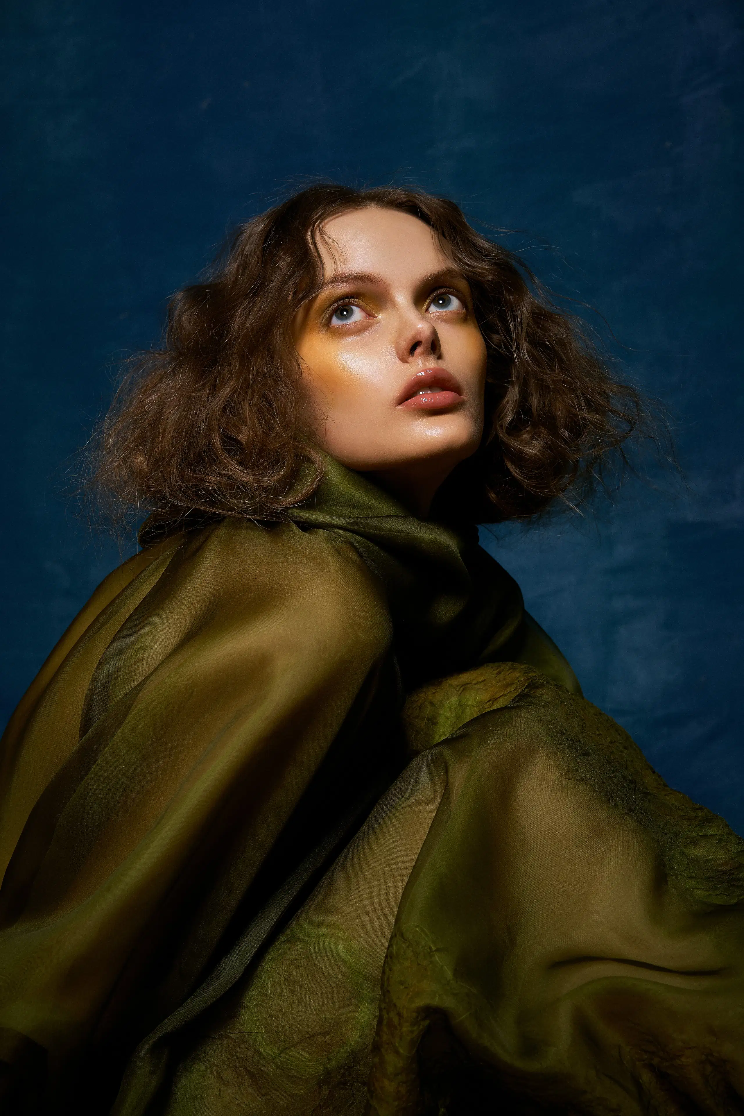 A fashion portrait of a woman against a blue background. She has shoulder length, wavy hair, is wearing a sheer green drapery, and looking up and off camera.