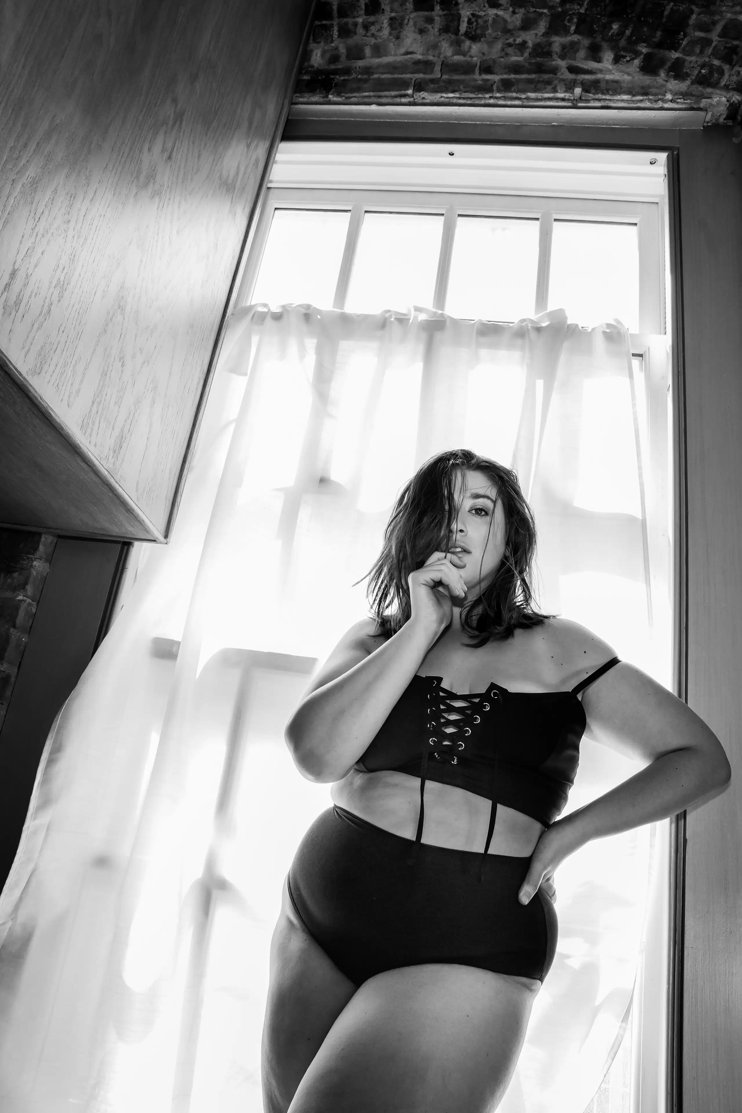 A black and white photograph of a woman wearing dark lingerie. She is standing in front of a long window with white sheer curtains in front of it. She has shoulder length hair that is falling into her face. One of her hands is holding some of her hair, and the other is on her hip. She is looking at the camera.