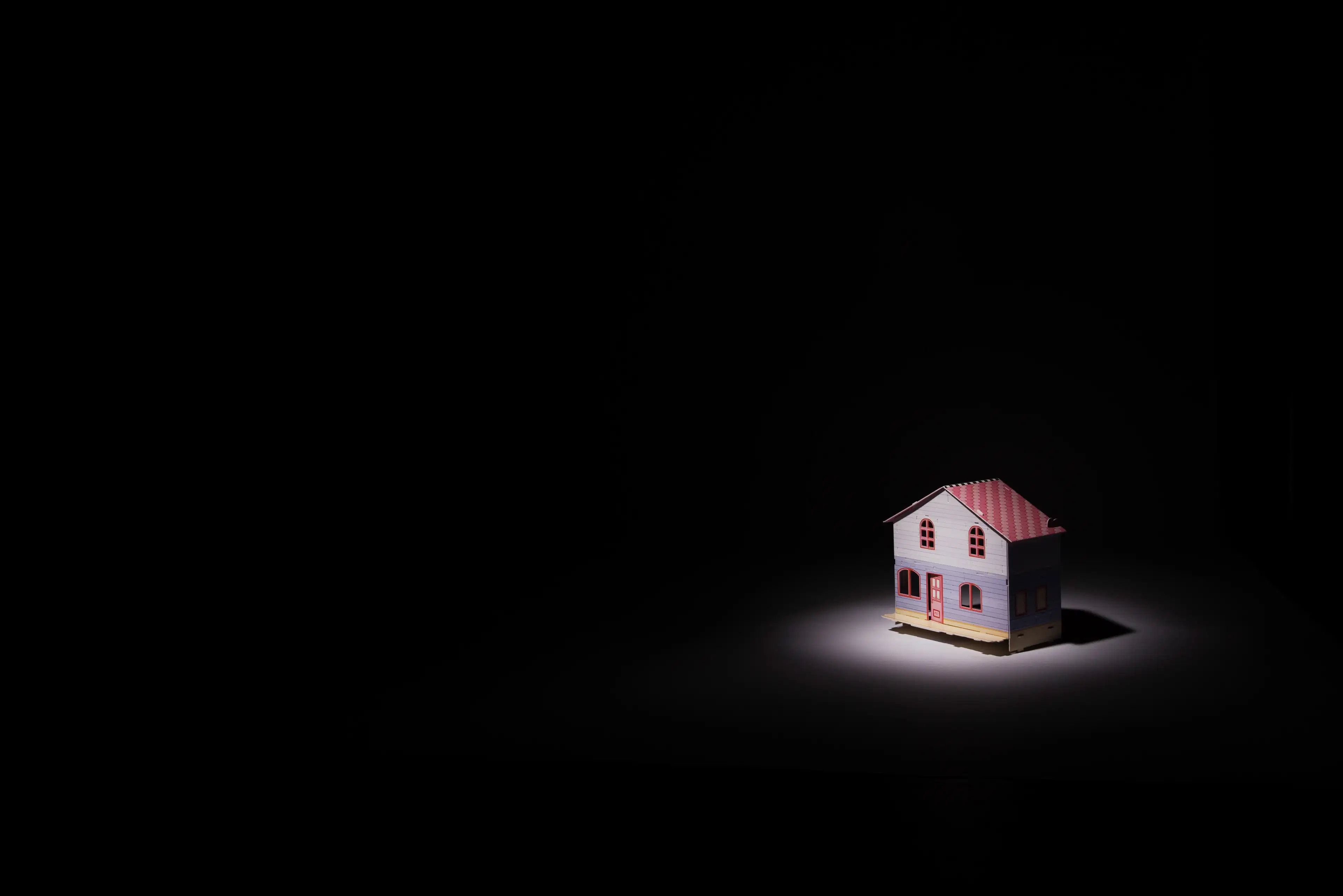 A still-life photograph of a dollhouse sitting on the floor. It is lit by a spotlight, the rest of the image in darkness.