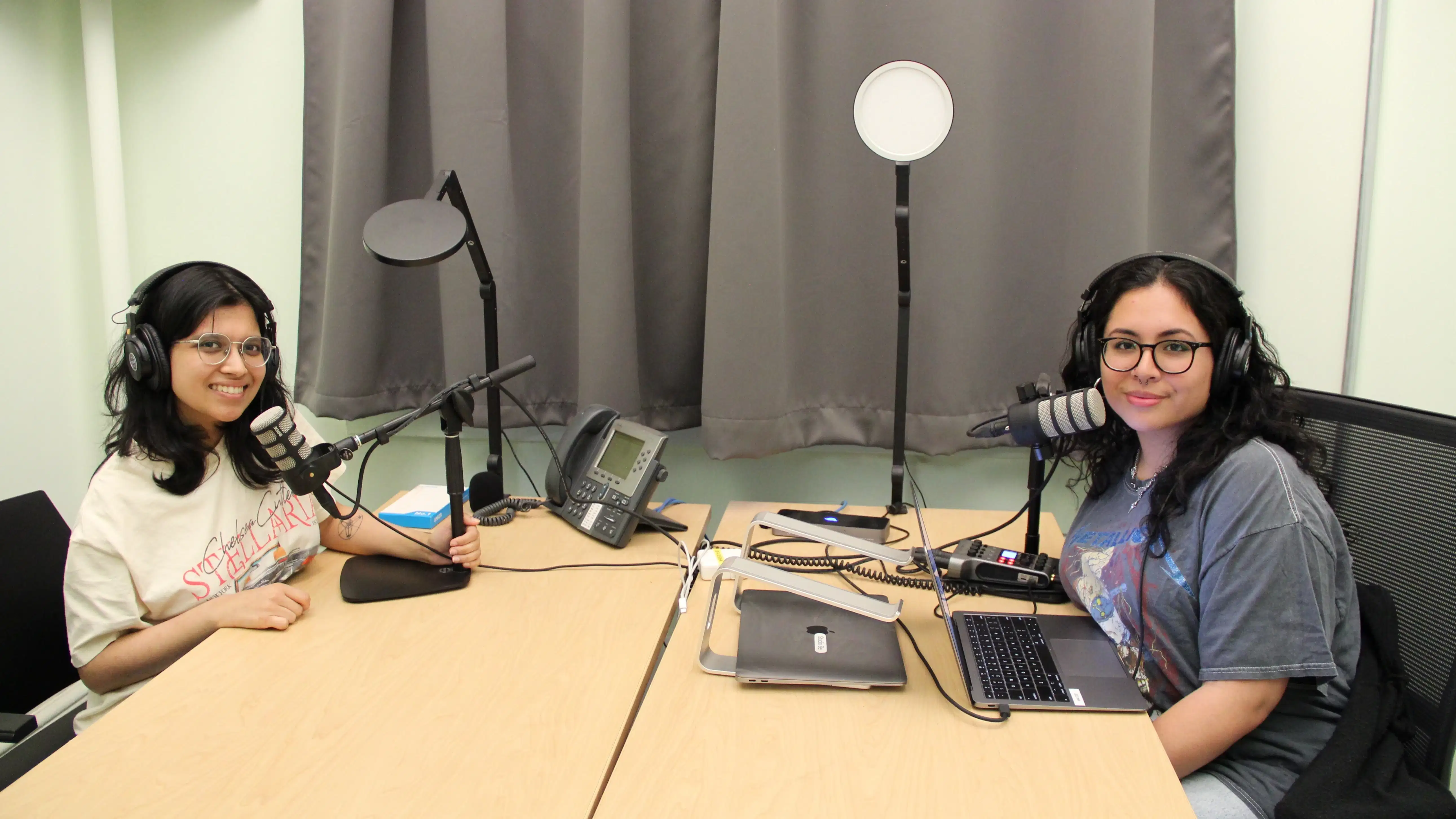 Two people sit at a long opposite one another smiling for the camera; they are using podcast production equipment such as microphones, laptops, headphones and lights.