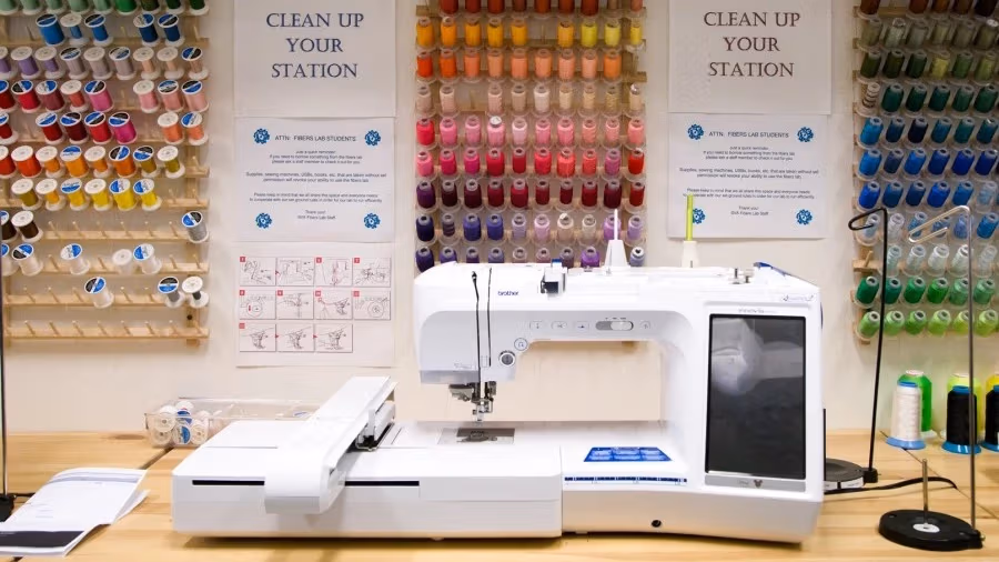 Fabric studio view of a professional grade sewing machine and various colored spools of thread hanging on the wall.