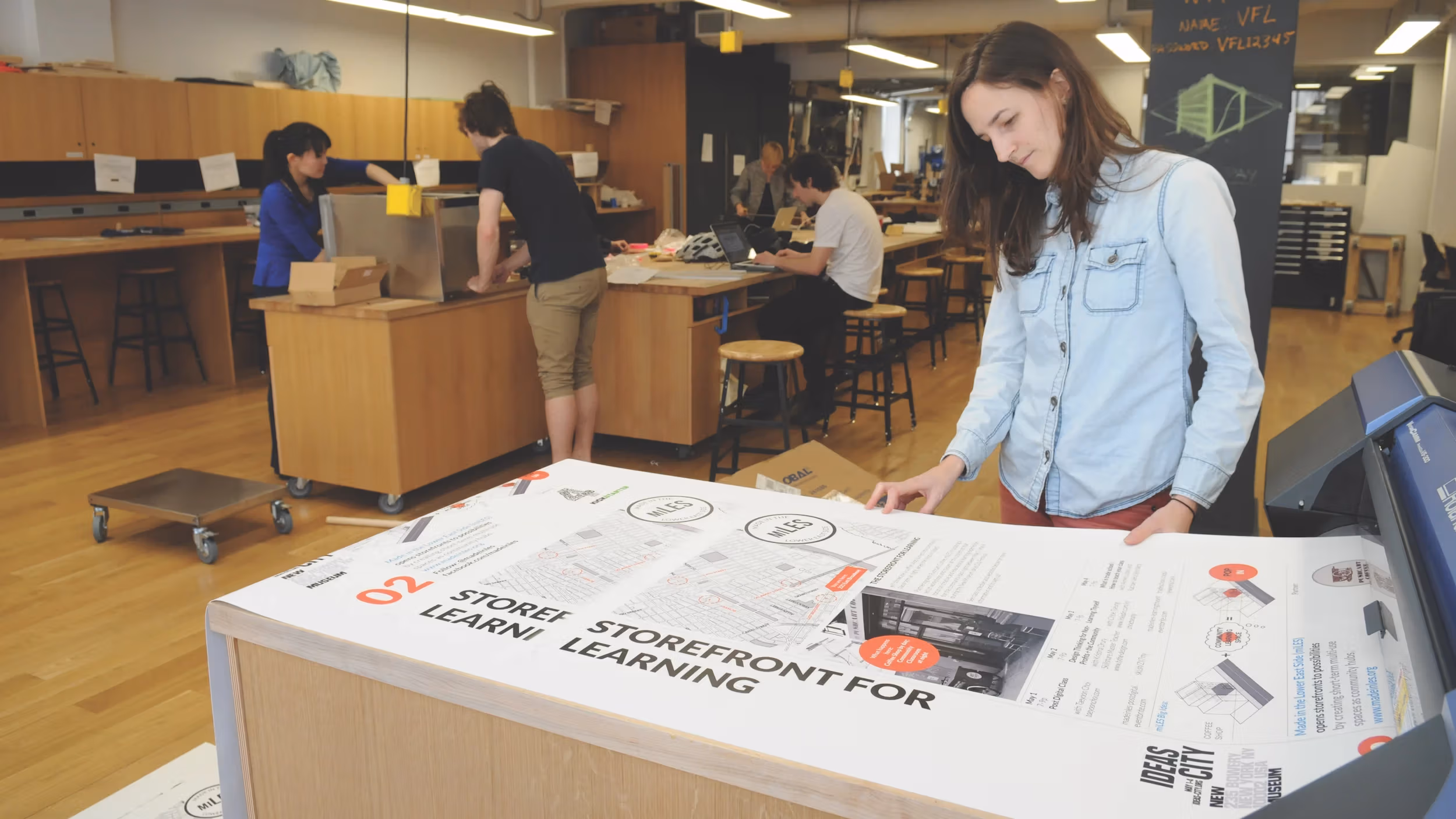 Wide view of a maker space with wooden floors, cabinetry, and workspaces. In the foreground, a student looks on as their project is being printed from a large printer.