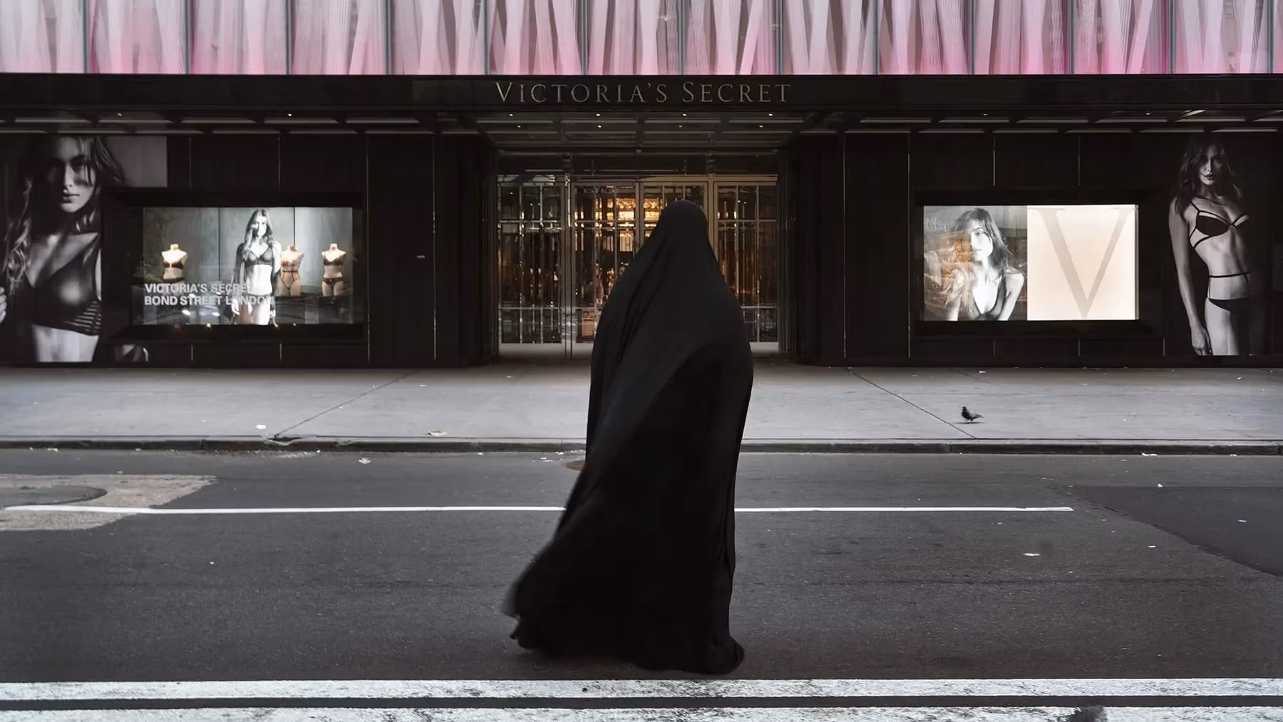 A color photograph of a figure enveloped in a flowing black garment walks across a city street, directly in front of a large Victoria's Secret store. The storefront features large, illuminated advertisements of models in lingerie on either side of its main entrance.