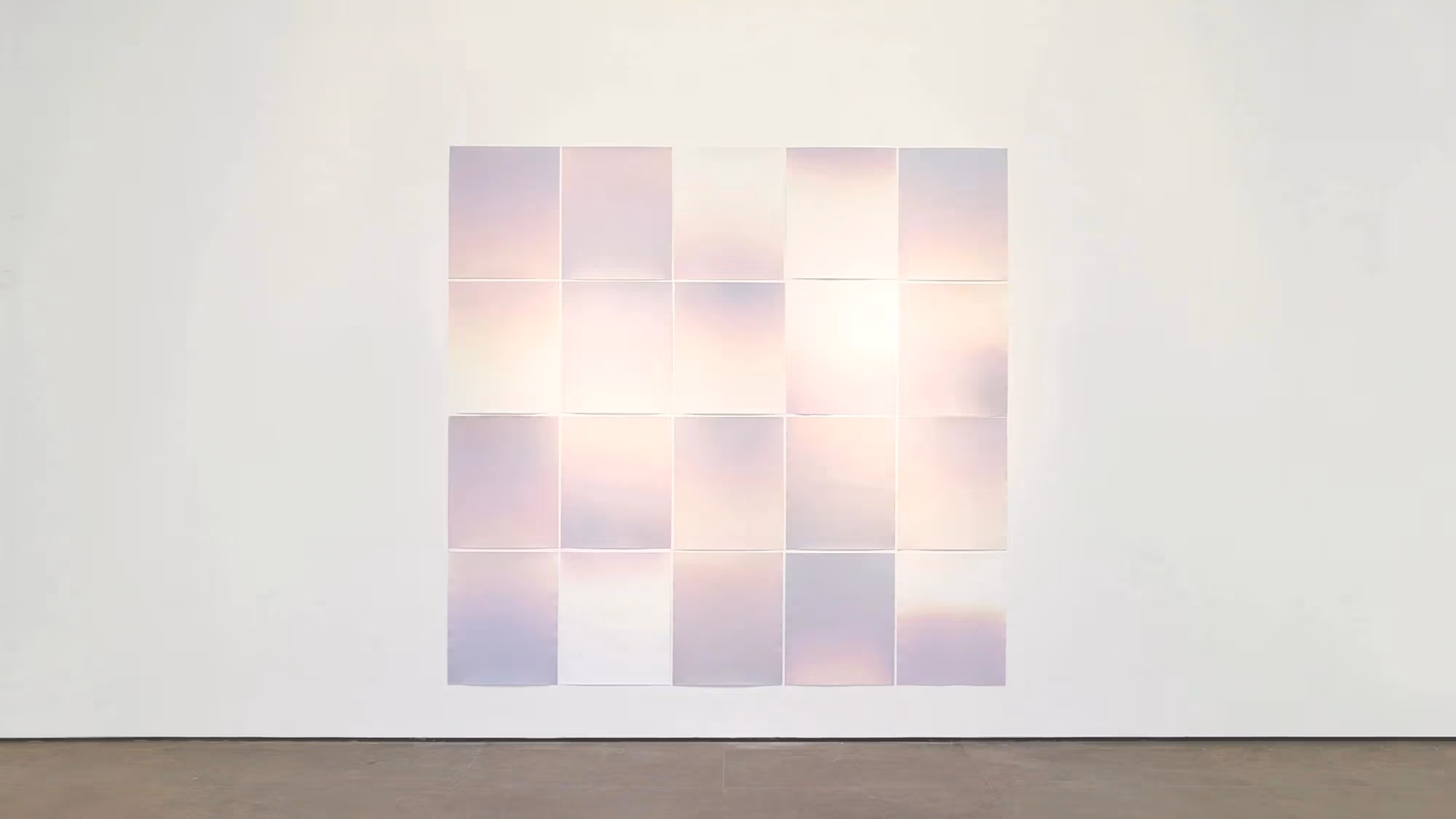 A large grid of soft, pastel-colored photographic prints is arranged on a white gallery wall. Each print features gentle gradients of pink, lavender, and light cream, creating a luminous, atmospheric effect. The grid forms a rectangular composition that glows subtly against the minimalist space