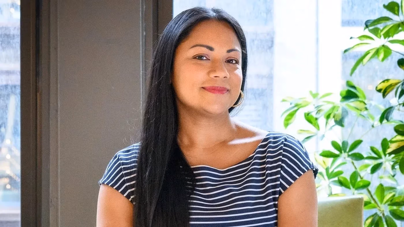 A person with long dark hair wearing a navy and white striped dress sits on a green chair, hands clasped, in a bright room with large windows and a leafy plant.
