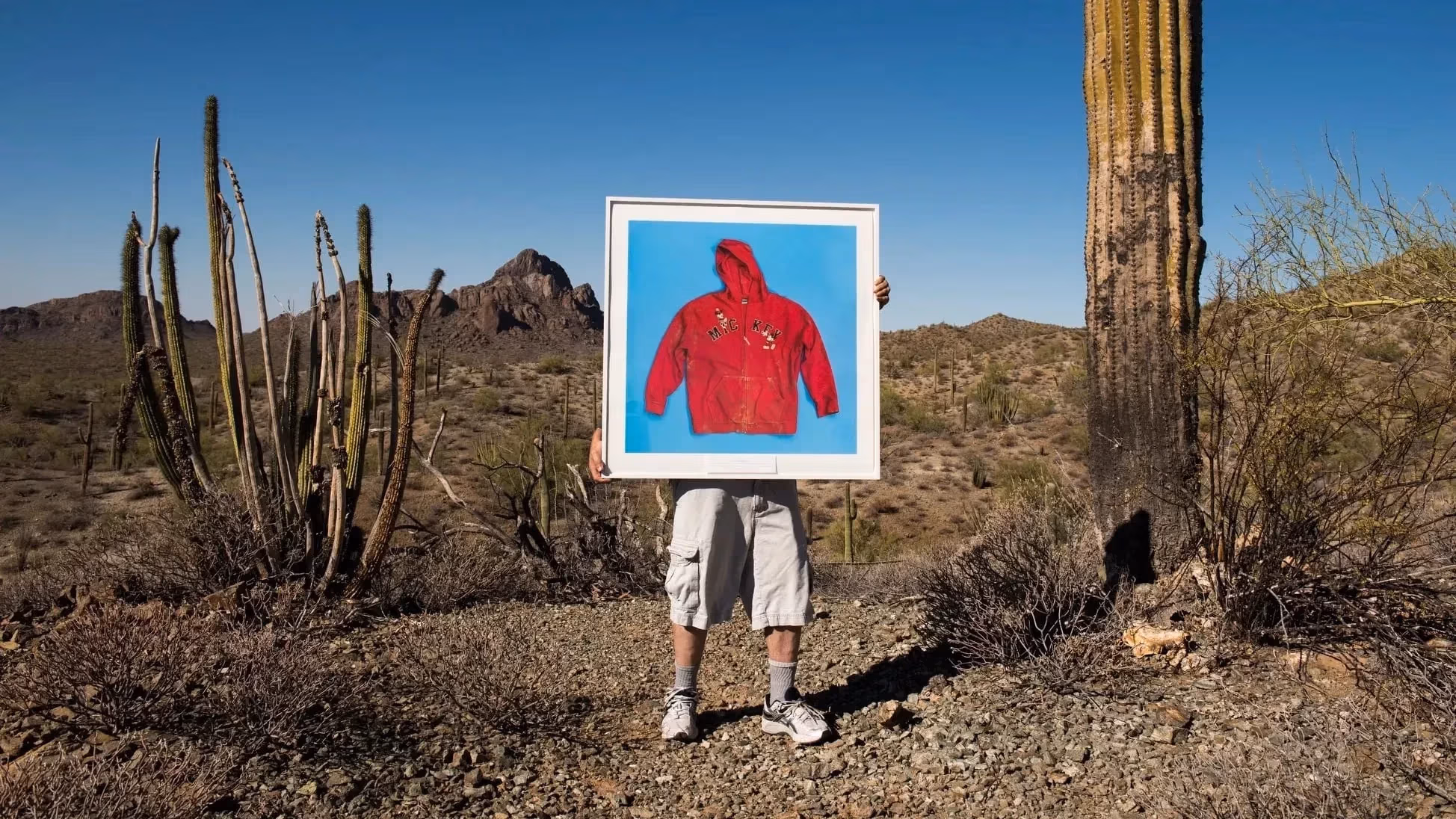 Photographer Tom Kiefer near his studio in in Ajo, Arizona, in the desert with a huge cactus next to him. He is holding up one of his framed photos of a red hooded sweater on a blue background. The letters on the sweater say MICKEY.