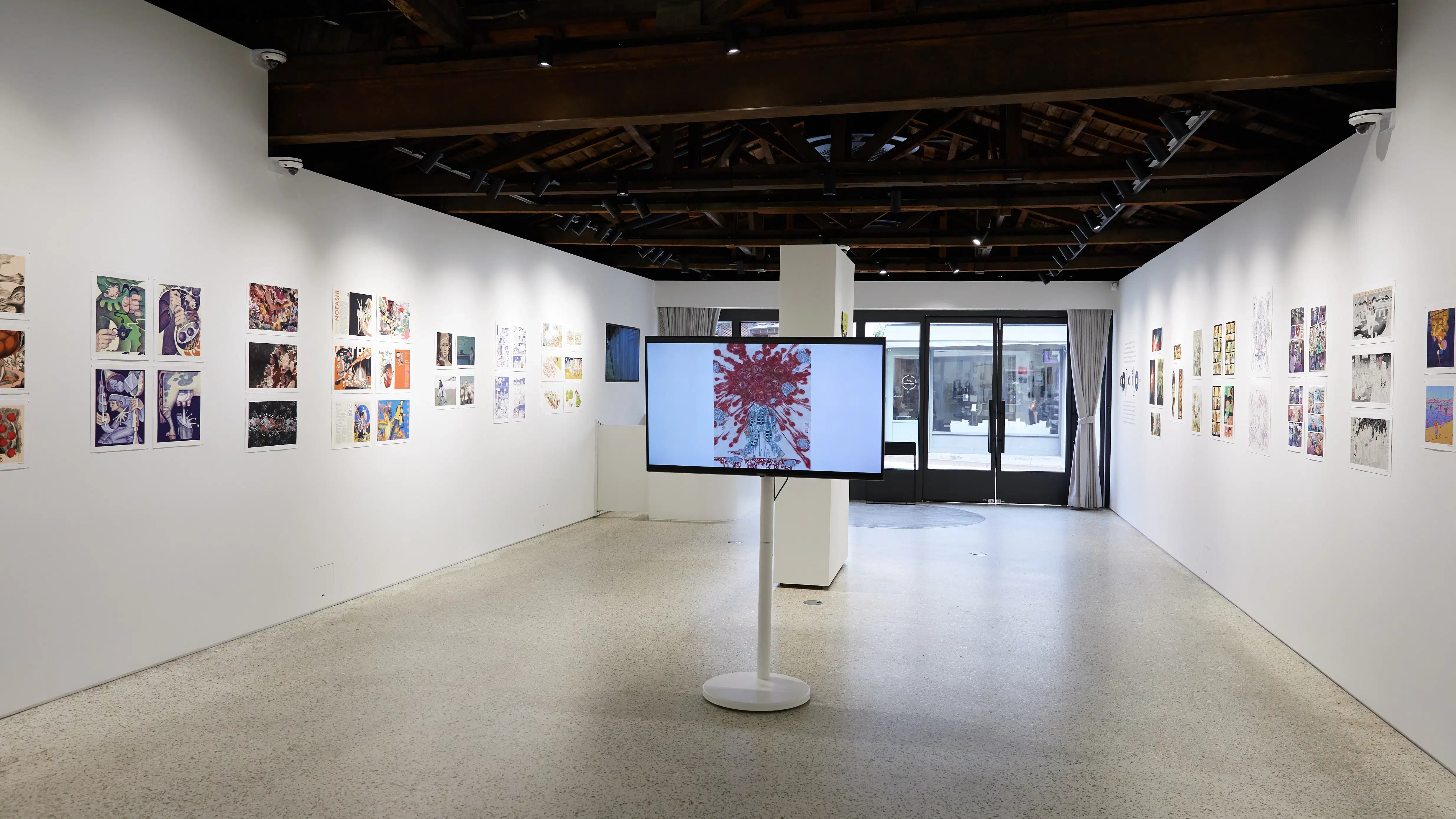 Gallery installation displaying colorful comic and illustration-style artworks arranged linearly along both walls. A large monitor on a white pedestal at the center shows a figure with red abstract patterns. The space features an open ceiling with exposed dark wooden beams, track lighting, and natural light filtering through glass doors at the far end.