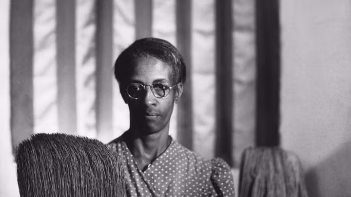 A Black woman stands in front of the American flag holding a broom and a mop.