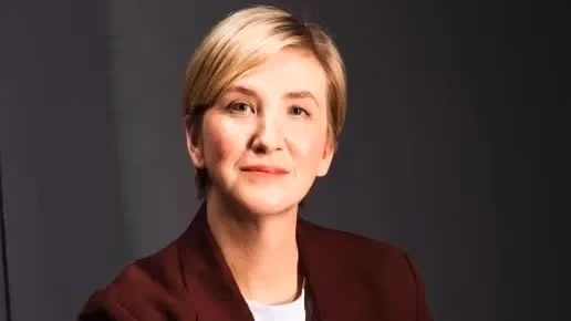 a person with short blonde hair sits in a red blazer