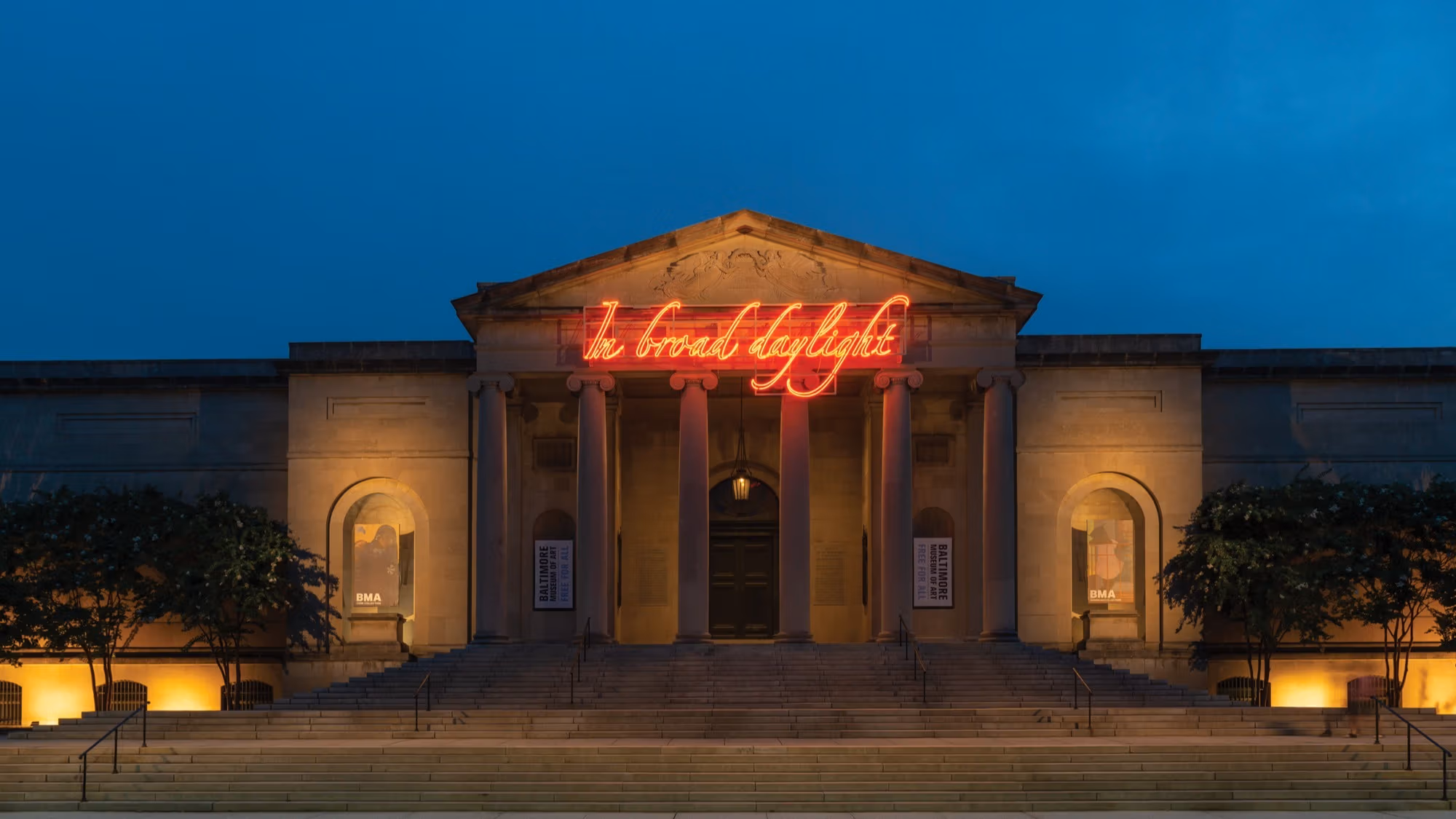 Installation view of Tavares Strachan’s work In Broad Daylight, installed at the Baltimore Museum of Art in 2018. A neoclassical style building at dusk features neon lights that read "In Broad Daylight" in reddish orange.