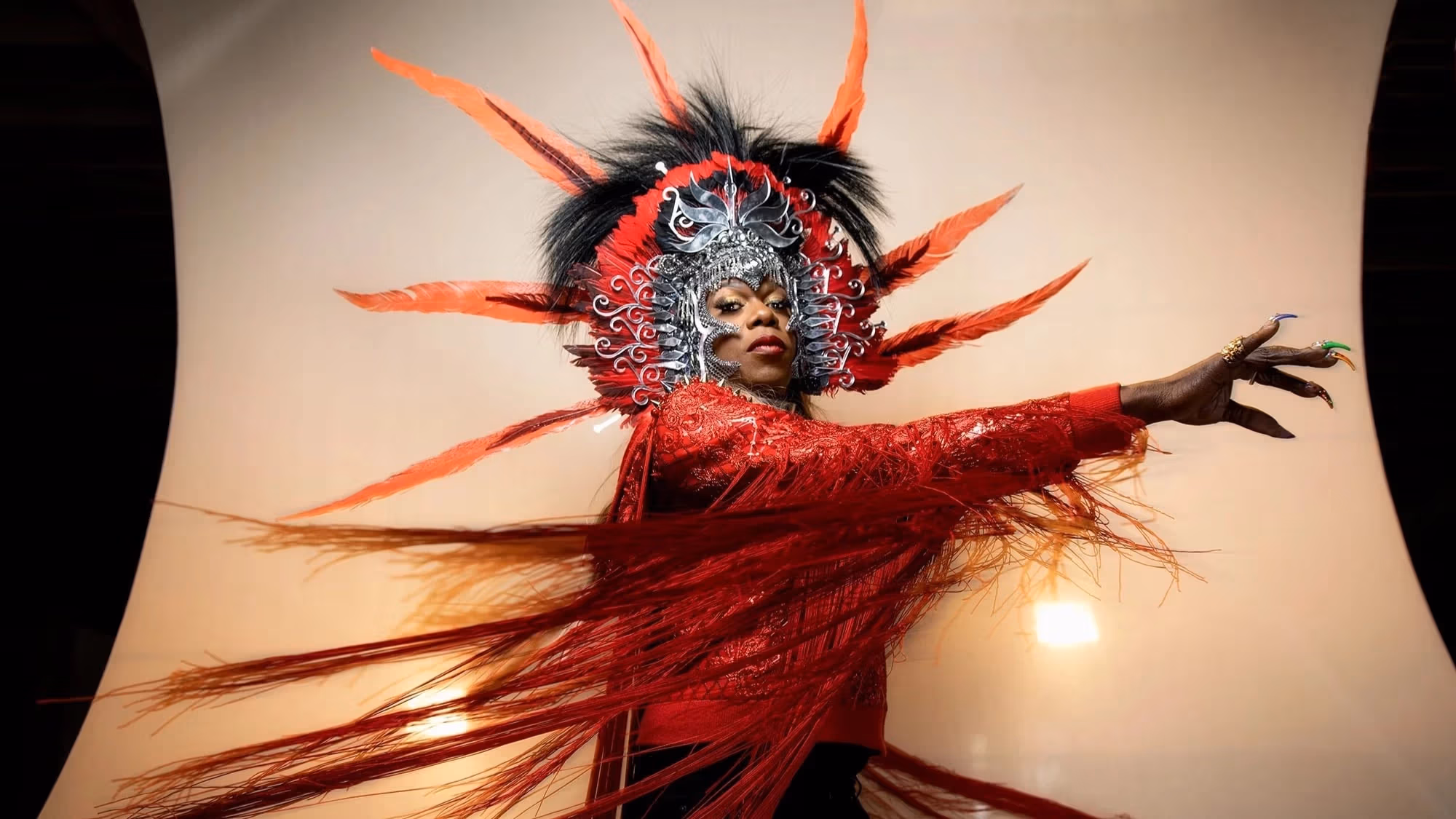 A studio portrait of a person in an elaborate headdress and red shirt.