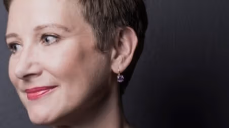 A headshot-style photograph of a person who is posed in front of a blank background, looking off to the left and smiling.