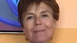 a person with short brown hair and brown eyes smiles in front of a beige wall 