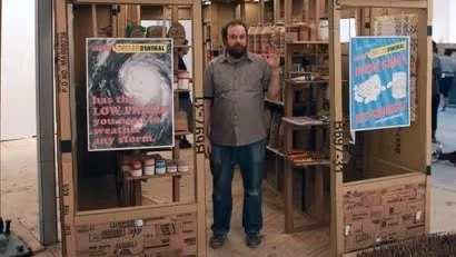 A man standing in a cardboard rendition of popular dollar stores. He is standing in the middle of the "doorway" wearing a button-up shirt and blue jeans.