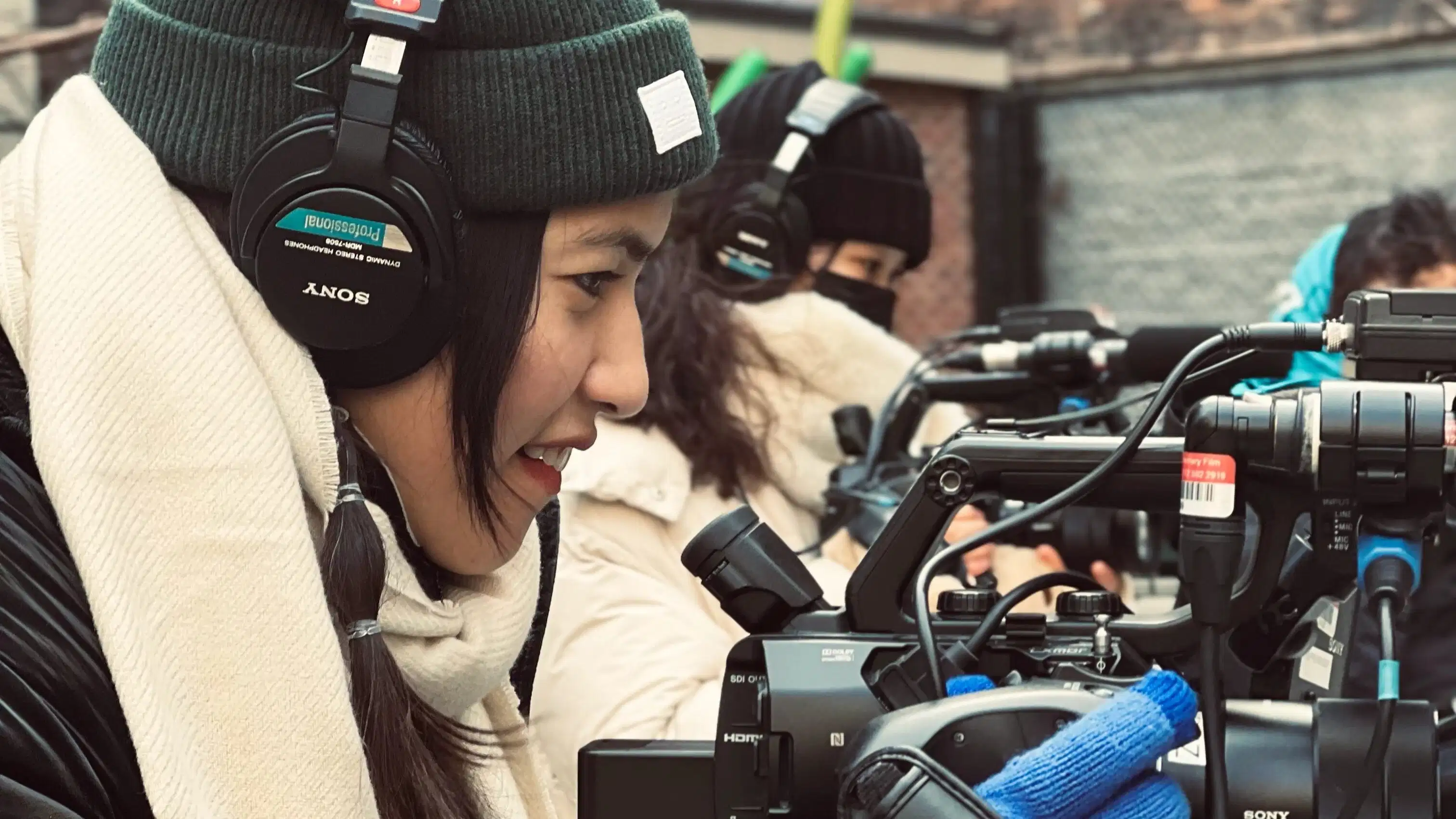 a woman films outside, wearing hat, gloves & headphones and in profile facing right. She looks into her camera. Another woman in a similar position is seen behind her.