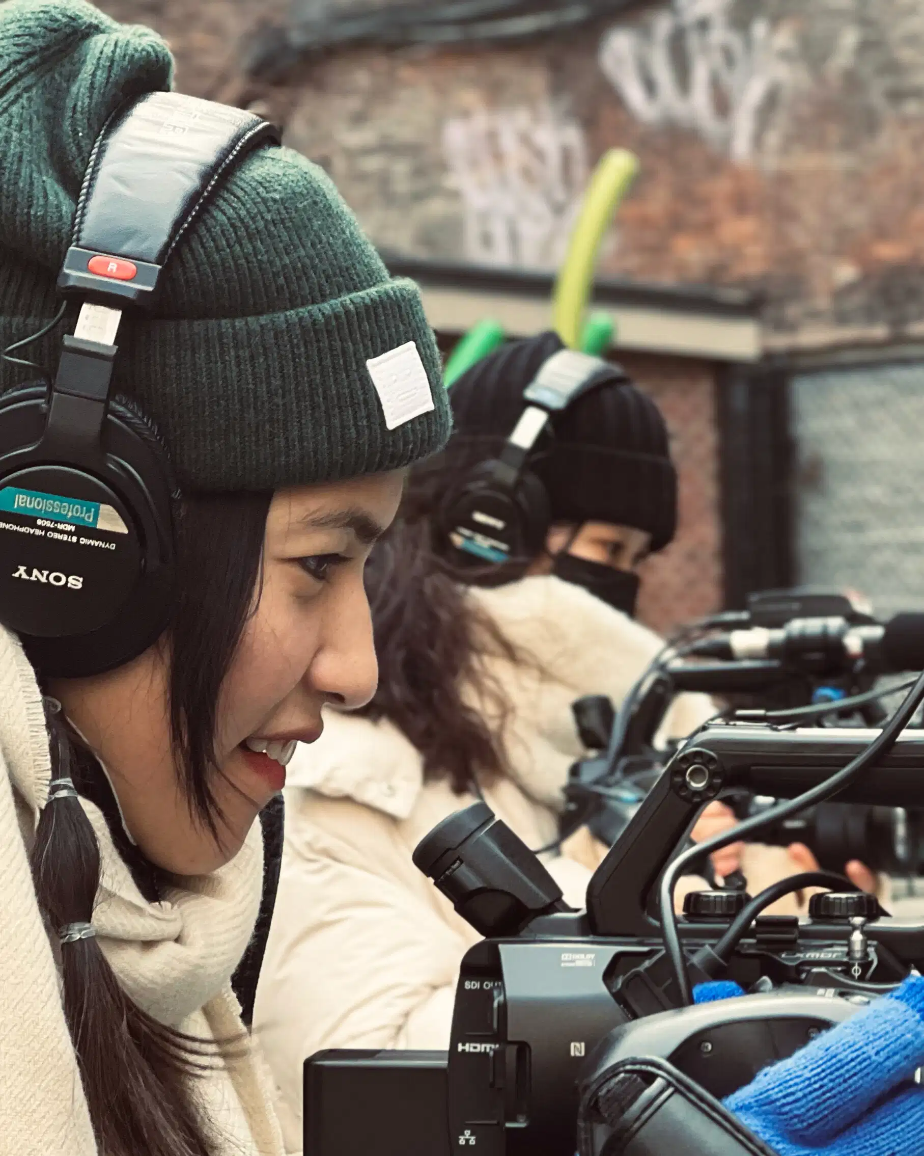 a woman films outside, wearing hat, gloves & headphones and in profile facing right. She looks into her camera. Another woman in a similar position is seen behind her.