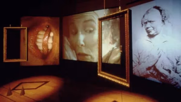 An image of a dark room looking at a diagonal angled wall with several spot lit frames hanging. The frames contain close up portraits. 