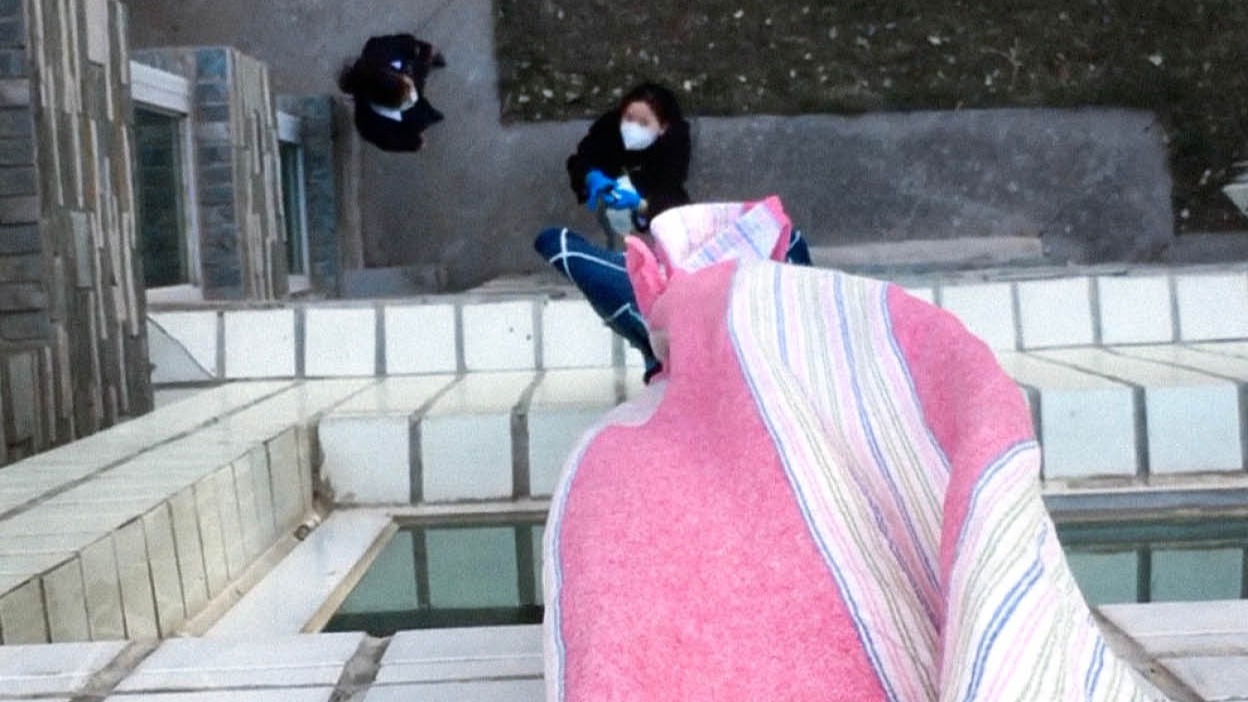  In the photograph, the viewpoint appears to be from above and looks down onto two figures. There is a pink and white stripped fabric that is hanging down the side of the building.