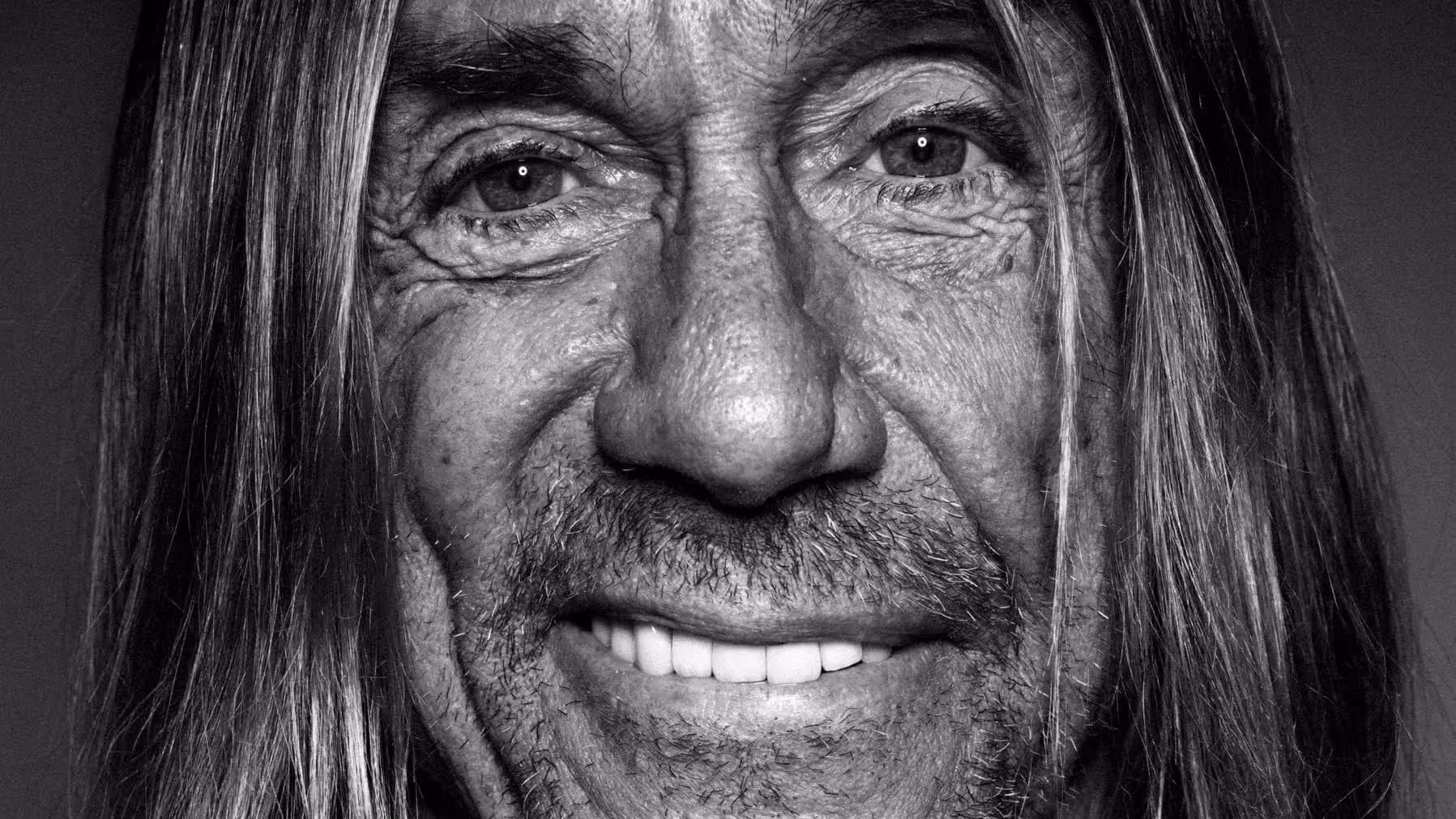 A black and white photo portrait of the singer Iggy Pop, who is looking straight into the camera with a smile. He has long hair and light skin, and is wearing a black cardigan over a gray t-shirt.