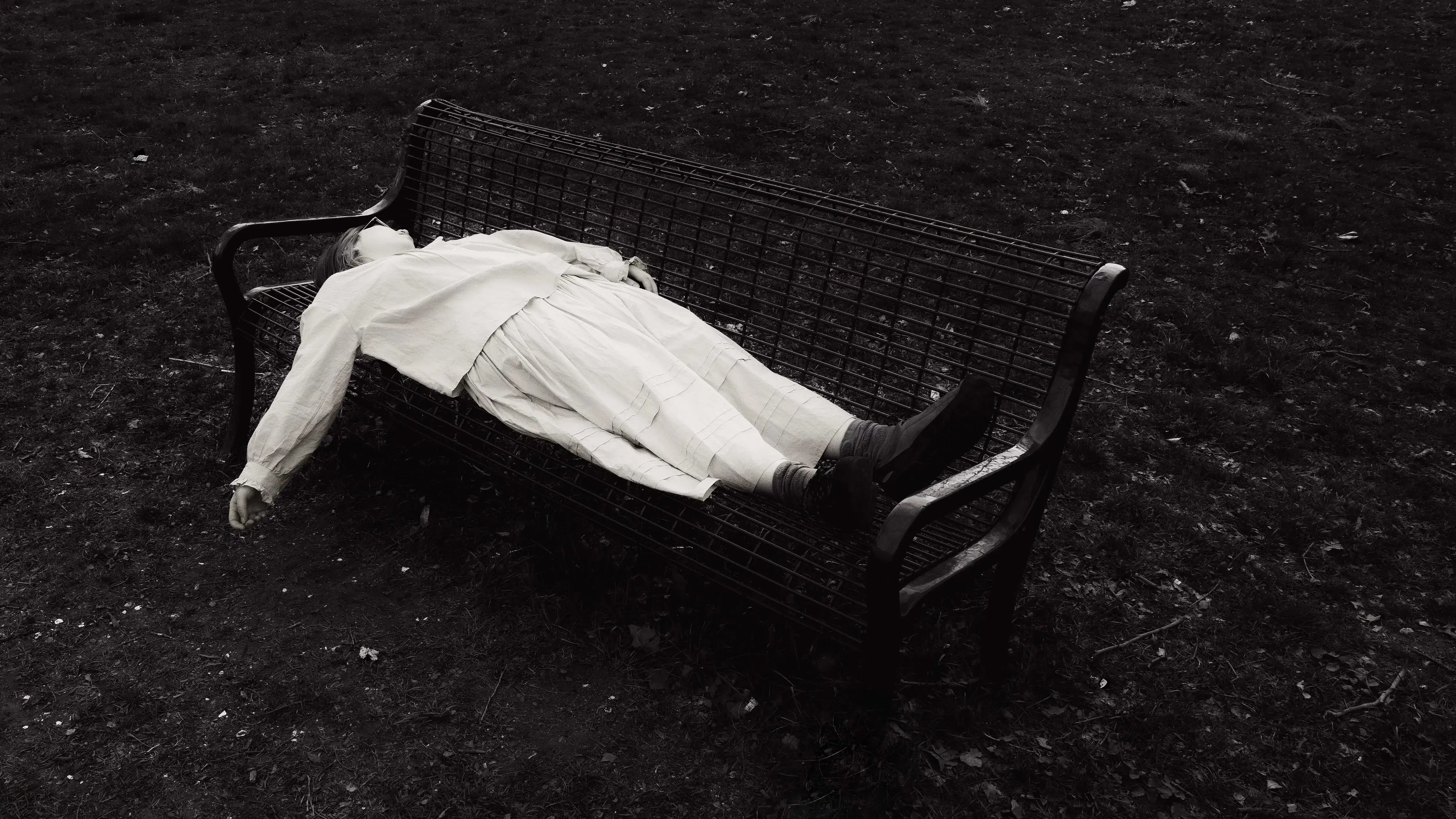 A black and white photograph of a girl lying on a bench, her hand hanging loosely, face obscured. The bench and her body are surrounded by chaotic bundles of grass.