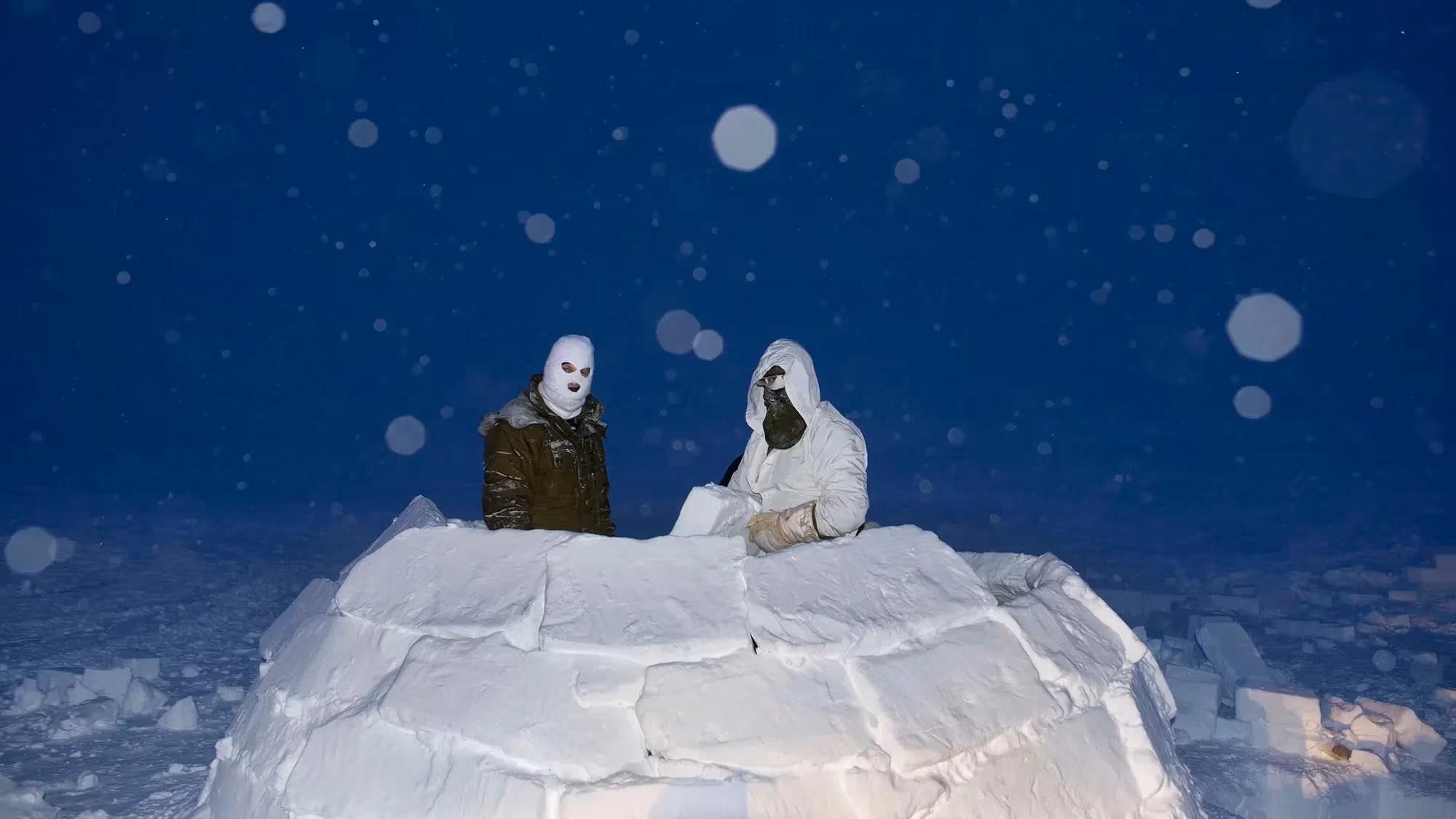A color photograph of Two soldiers, wearing masks in an igloo made of snow blocks, at dusk while it snows, lit with flash.