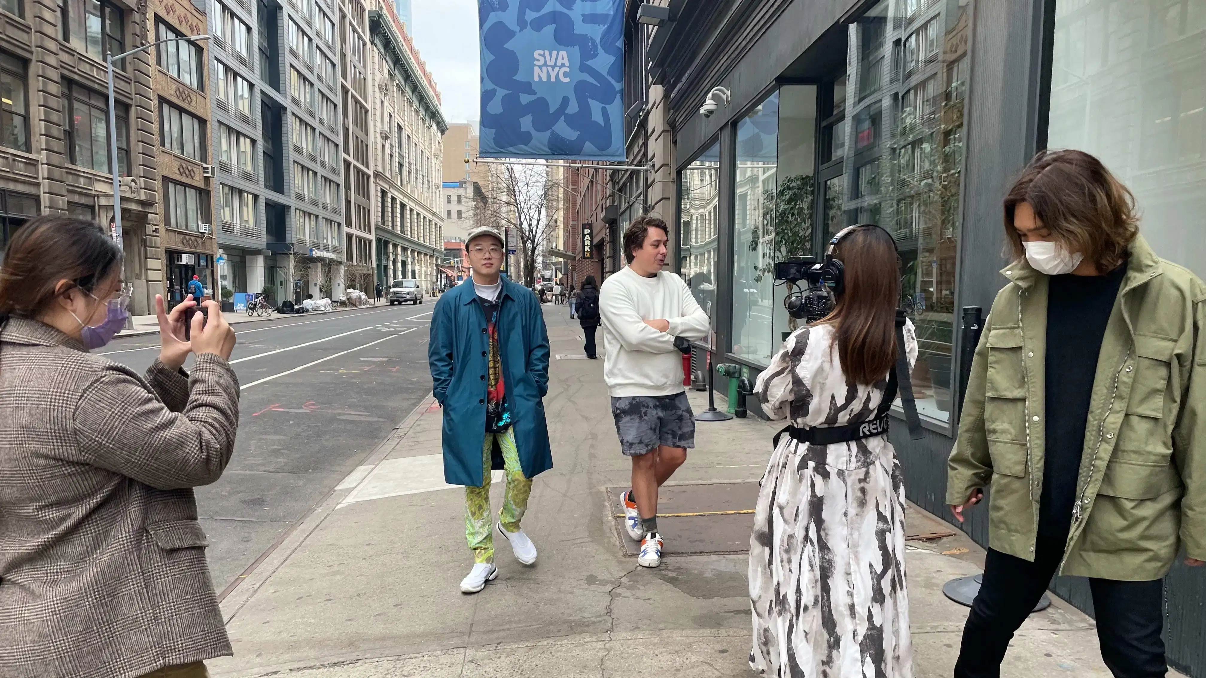 5 students walk towards the camera, 2 walk backwards holding cameras, filming another two. A blue SVA NYC banner flies in the background