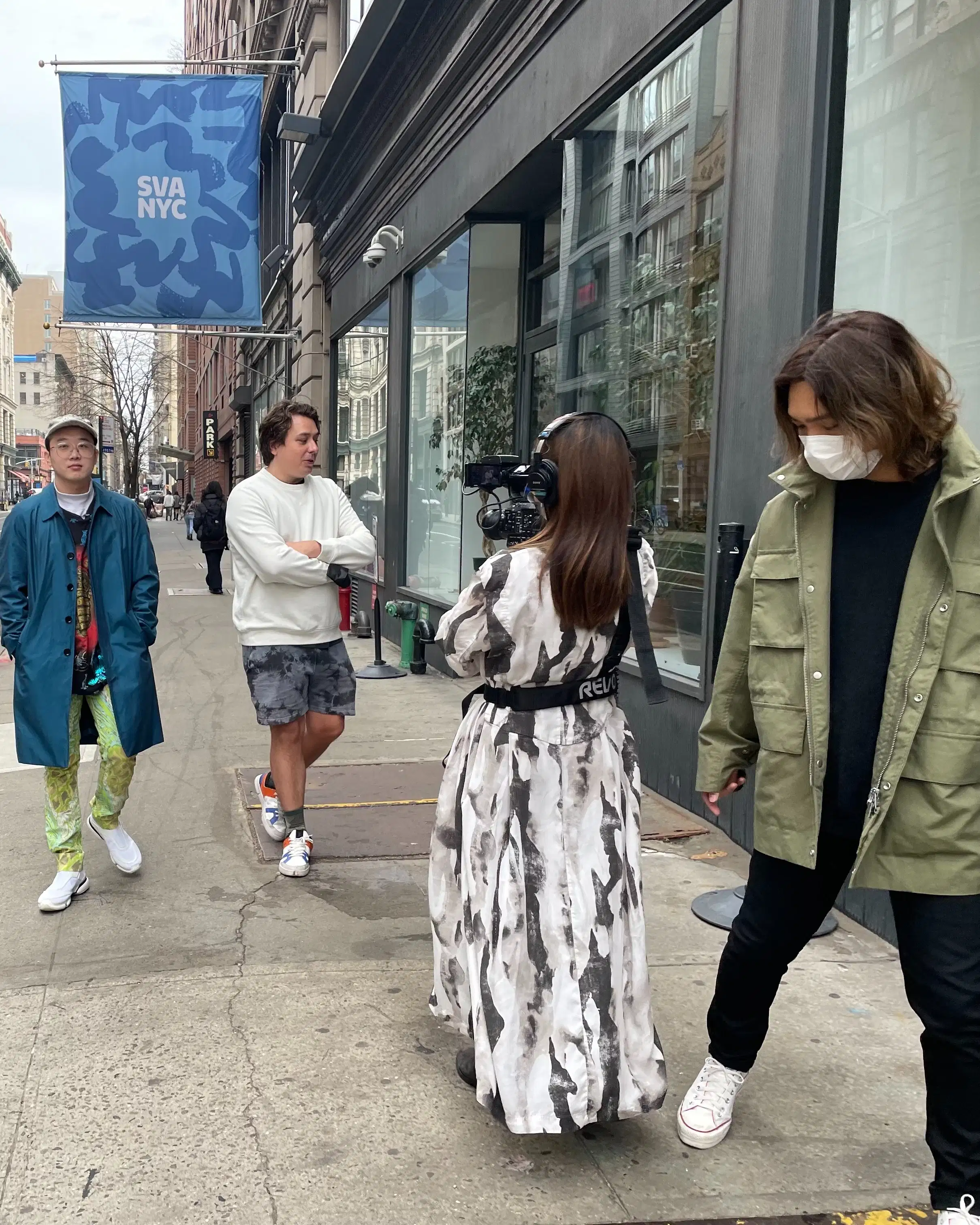5 students walk towards the camera, 2 walk backwards holding cameras, filming another two. A blue SVA NYC banner flies in the background