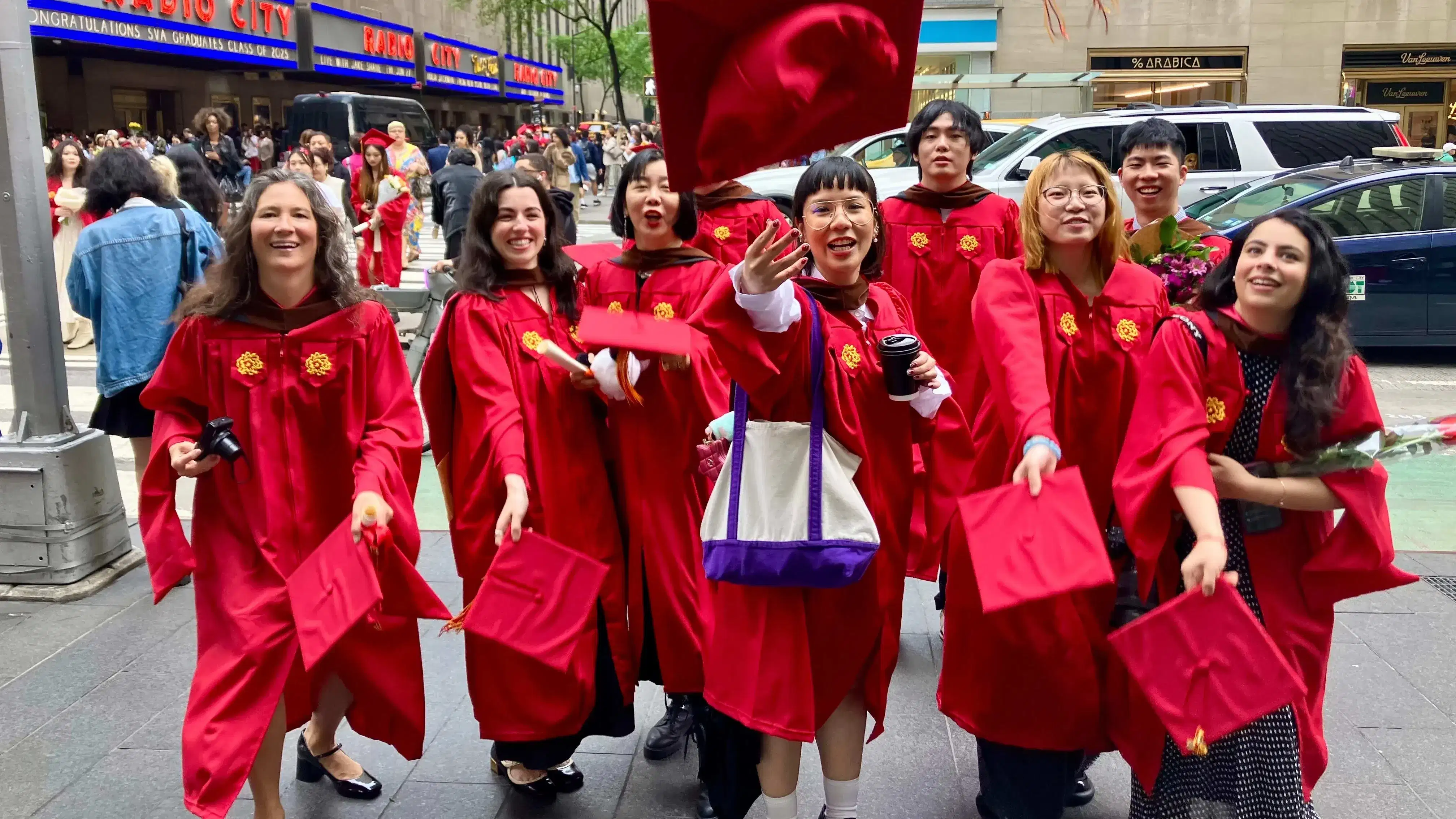 The Masters in Digital Photography graduating class of 2025, in the iconic SVA red graduation gowns, in front of the famed Radio City Music Hall where the commencement ceremony just took place. The students are threwing their caps in excitement. 