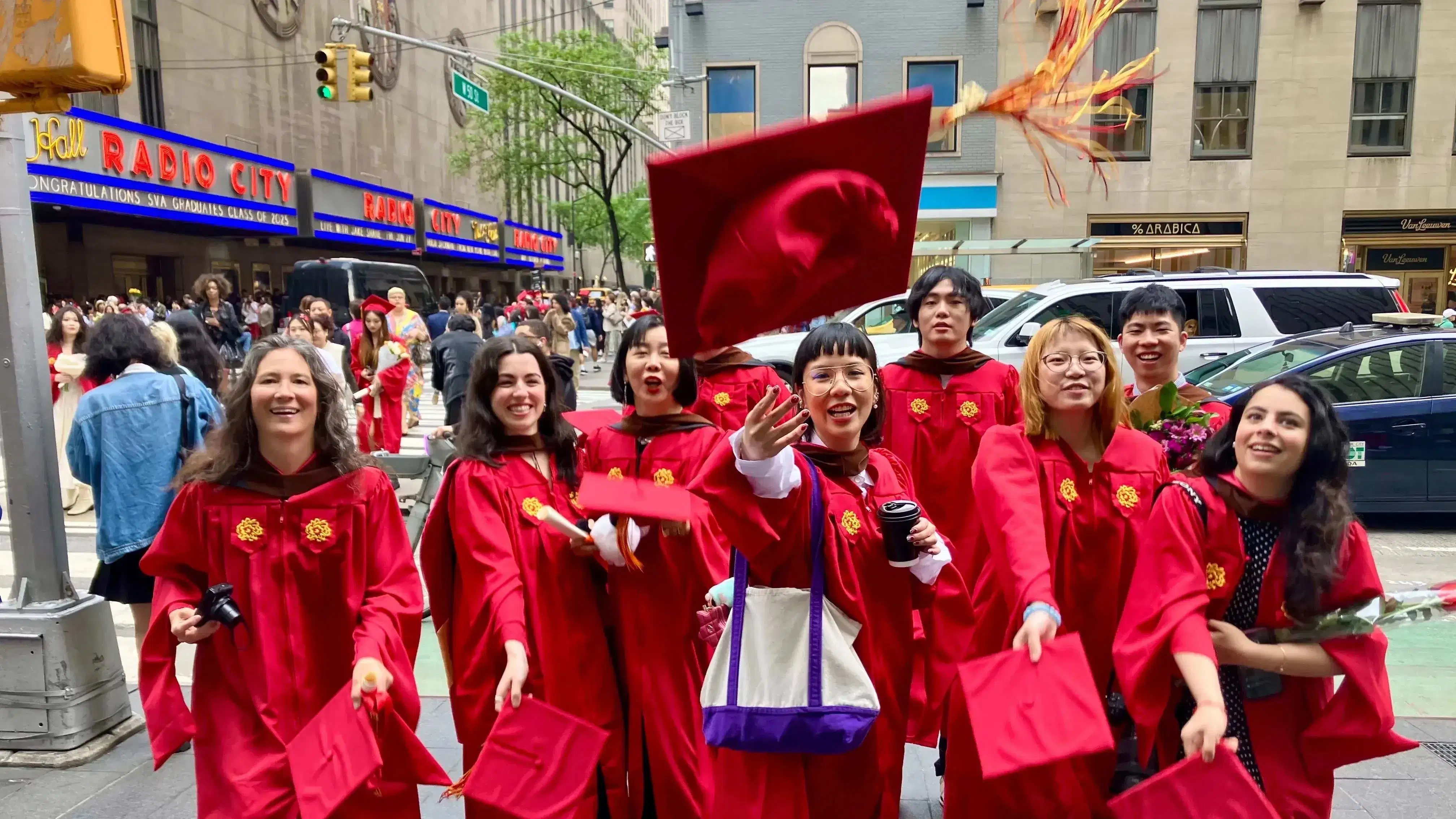 The Masters in Digital Photography graduating class of 2025, in the iconic SVA red graduation gowns, in front of the famed Radio City Music Hall where the commencement ceremony just took place. The students are throwing their caps in excitement. 