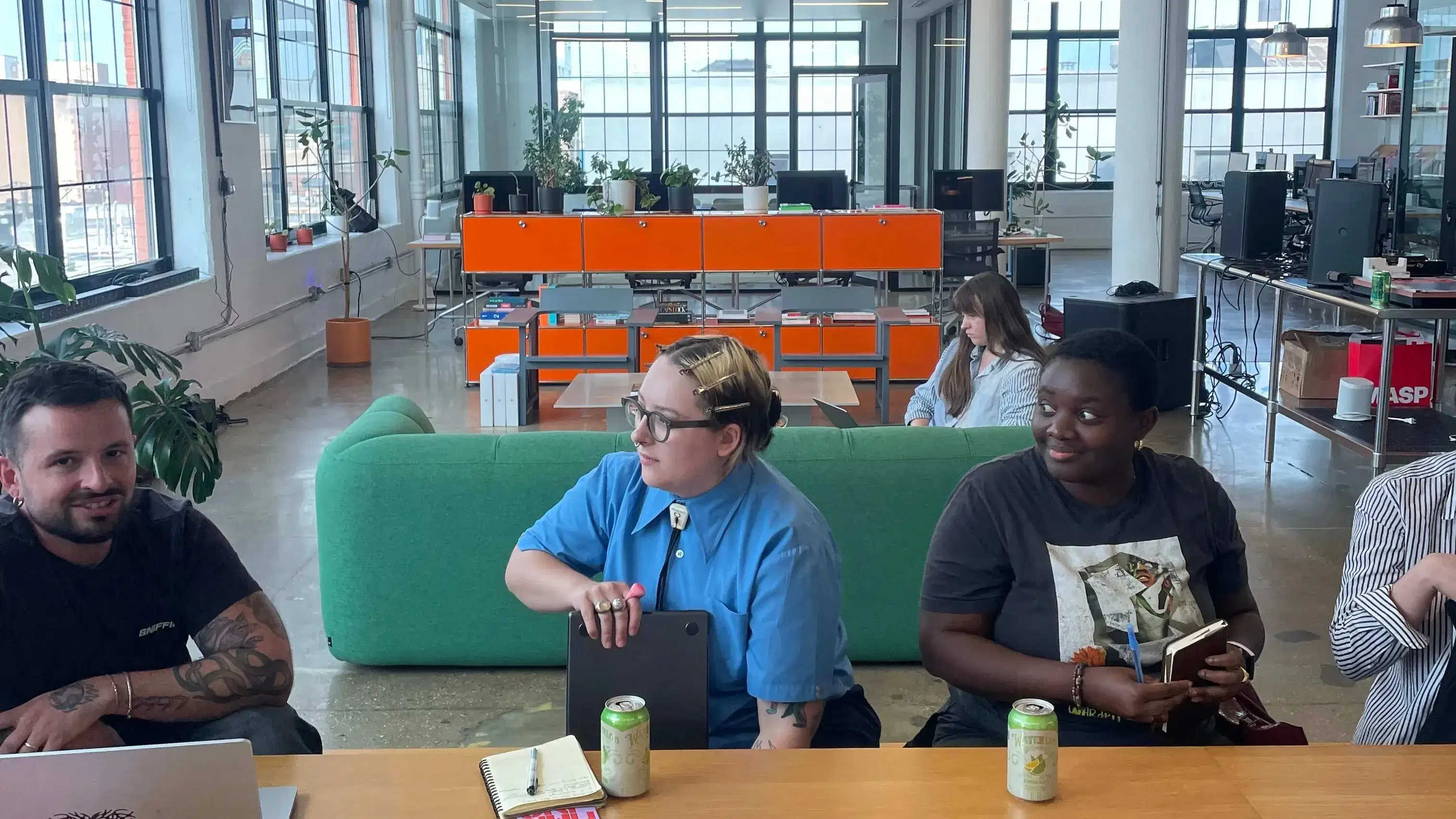 Alt text: Wide photo of a bright, industrial loft workspace with tall grid windows and exposed ducts. In the foreground, three people sit at a long wooden table—one with a laptop, others with notebooks and books—while soda cans and colorful magazines are spread across the tabletop. A green sofa and rows of desks and computers fill the background.