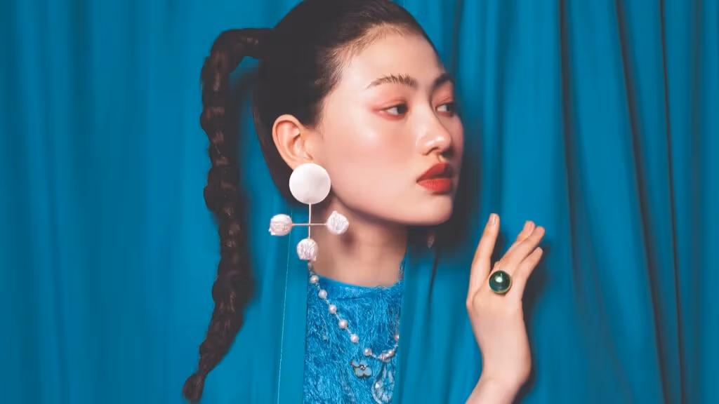 A woman peers out from behind a set of blue curtains. She is wearing a large white earring and a round green ring