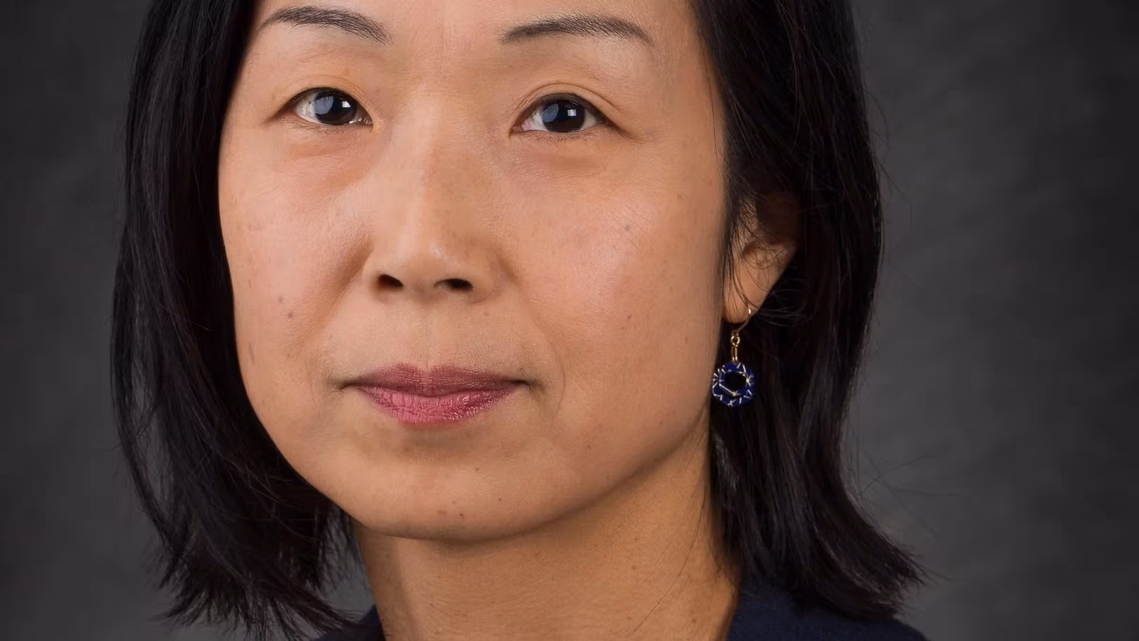A headshot of Midori Yoshimoto. Midori is wearing a stripped shirt with a navy sweater. She is set against a gray background and is looking at the camera..
