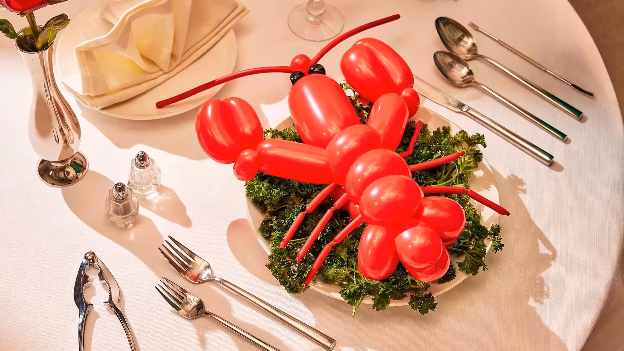 A photograph of a lobster shaped red balloon sitting on top of a bed of kale leaves on a dinner plate. Surrounding the scene is a dinner table fully set up with cutlery,  a dinner napkin folded into a fan, and a single rose in a tin vase.