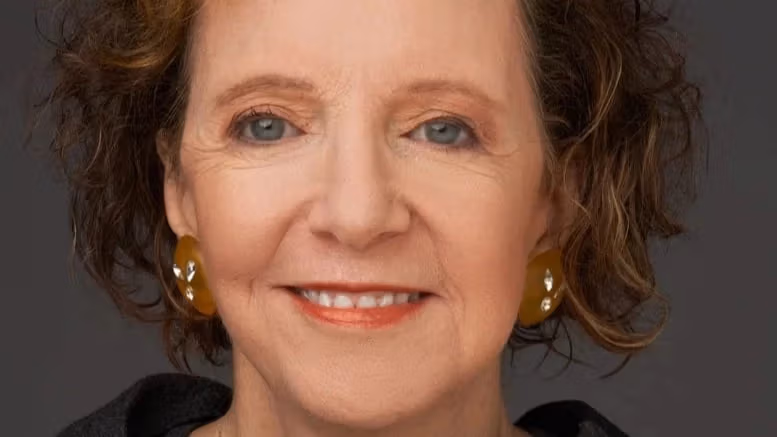 A photograph of Suzaan Boettger by Timothy Greenfield-Sanders. Suzaan is wearing a grey and black checkered sweater. She is smiling into the camera. The background is a solid grey color.