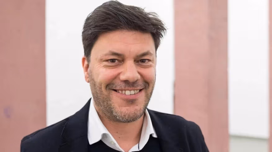 a smiling person with short brown hair and a black blazer on stands in front of a pink and white building exterior 