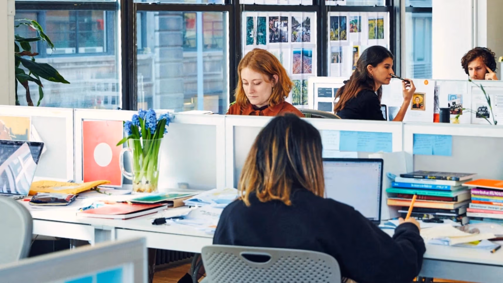 A person with orange hair sits at a row of desks working while other people in the room also work  