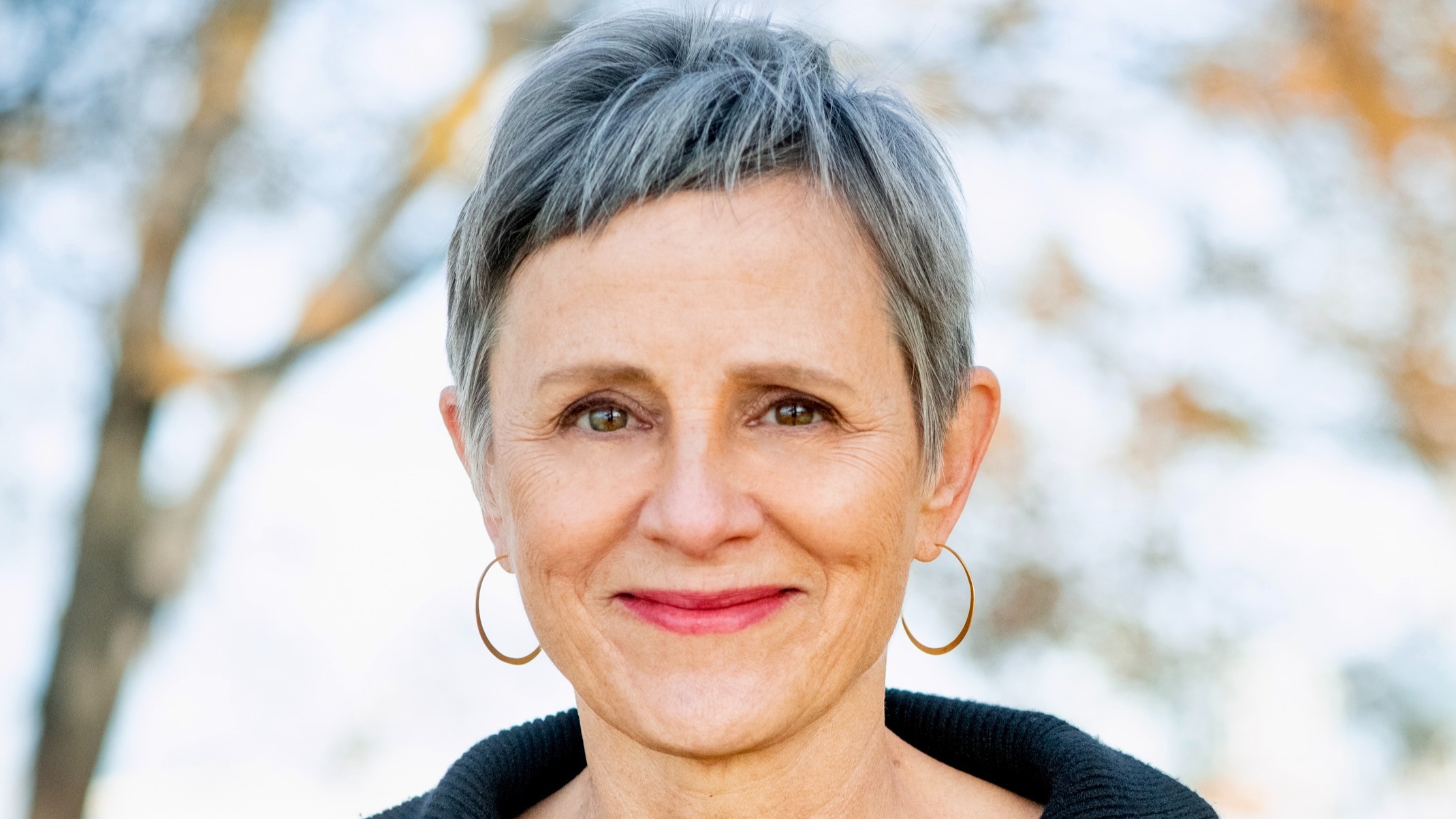 Susan Hapgood headshot. Susan is wearing a black sweater and has short grey and black hair. She is wearing thin hoop earrings and is smiling at the camera. In the background is a blurred natural scenery.