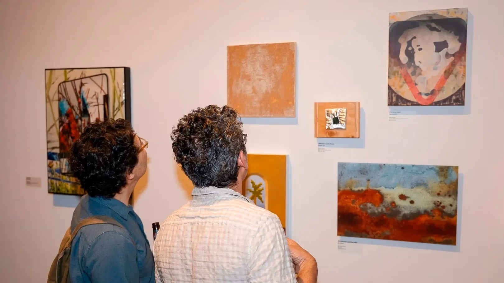 a person in a blue shirt and a person in a white shirt look at a wall of paintings