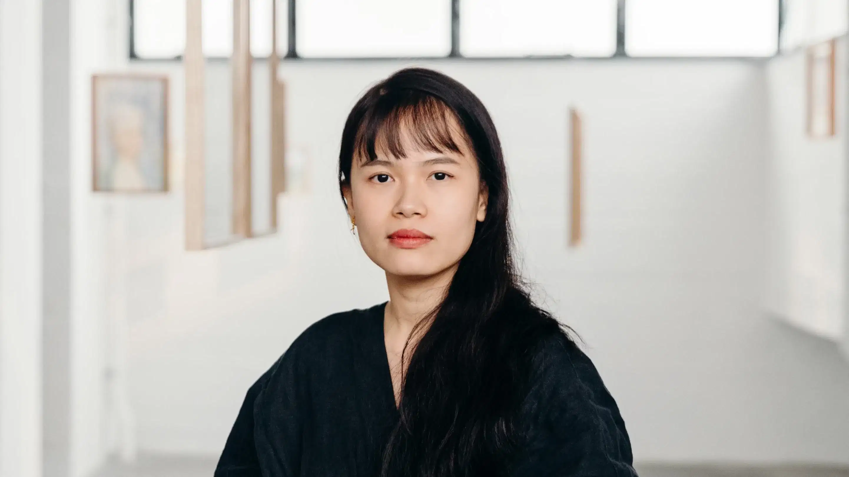 A photograph of a person wearing a black dress and standing in the center of the photograph looking at the camera. In the background the viewer can see some artworks hanging in a gallery.