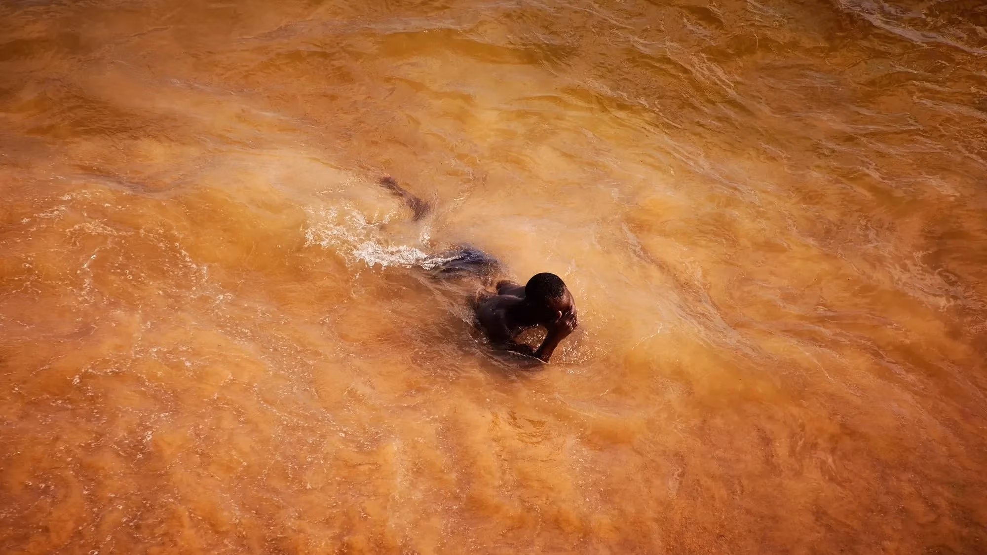 In this color photograph, a man emerges from the ocean, which has taken on a vibrant orange-ish cast in rural Saly, Senegal.
