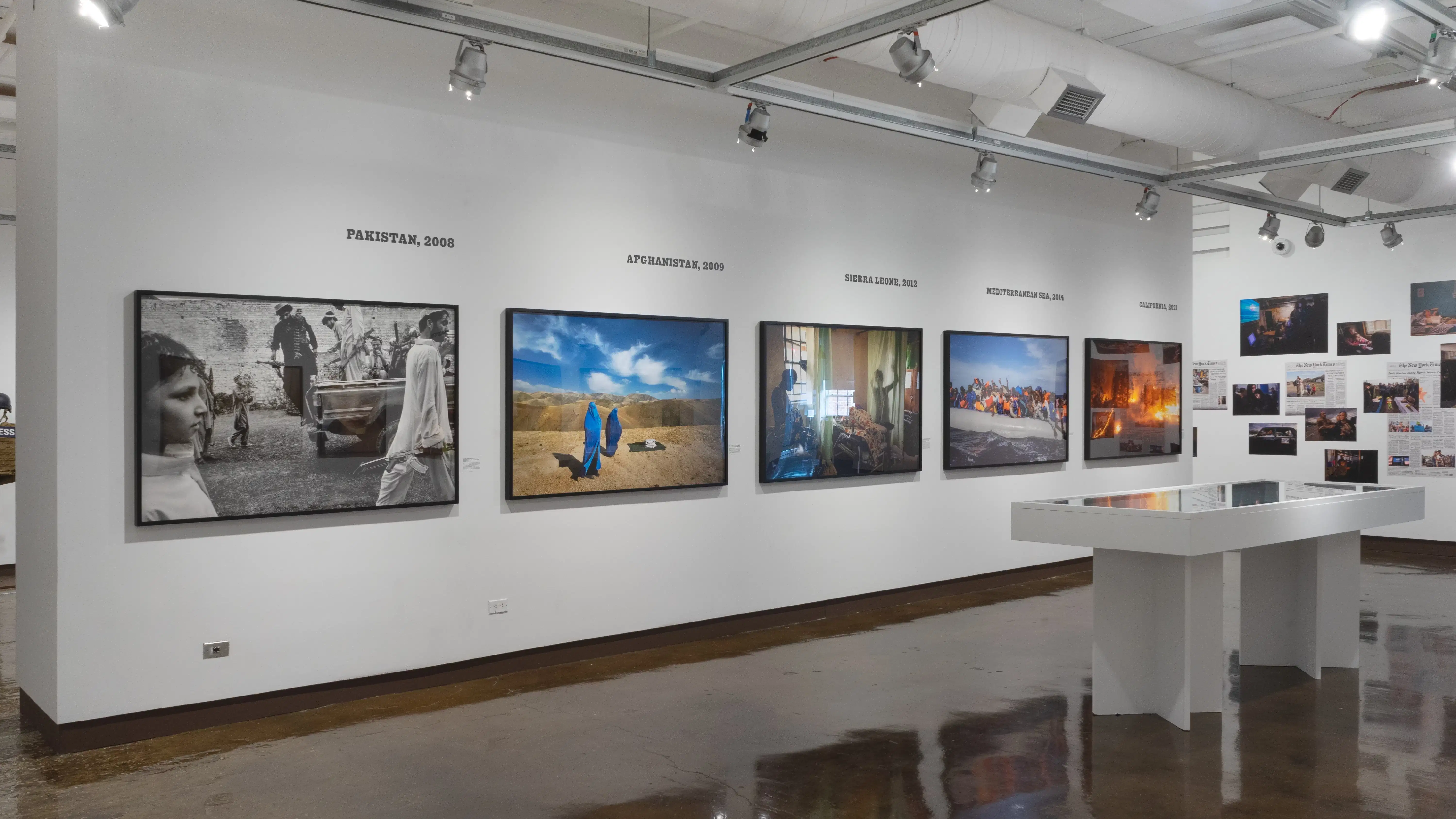 Photographs depicting war and conflict are hung across two gallery walls. In the center of the space is a large vitrine with ephemera inside. 