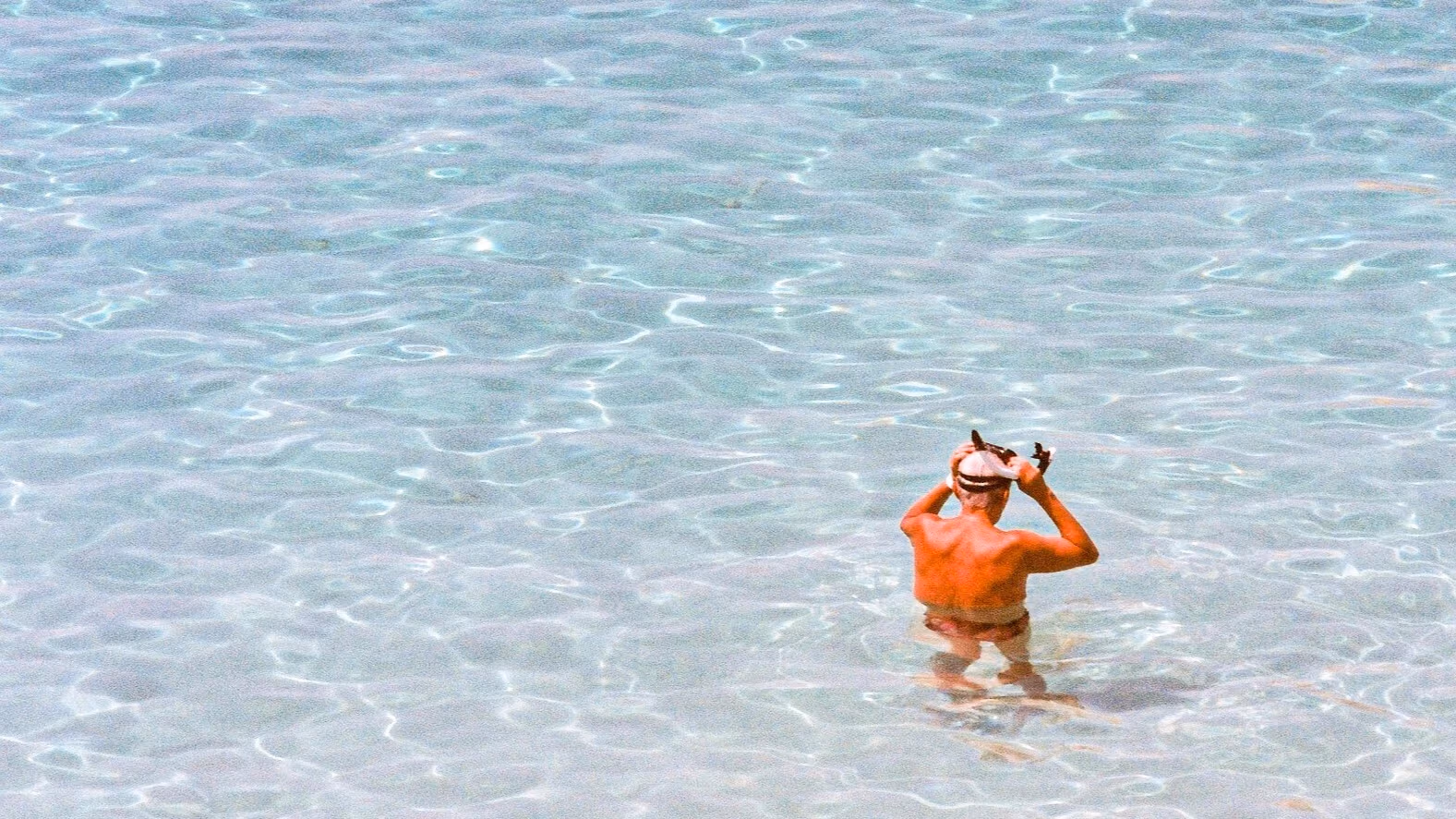 Sun drenched photo of an open sea landscape; in the bottom right corner, an older person is putting on goggles, seemingly about to go for a dip