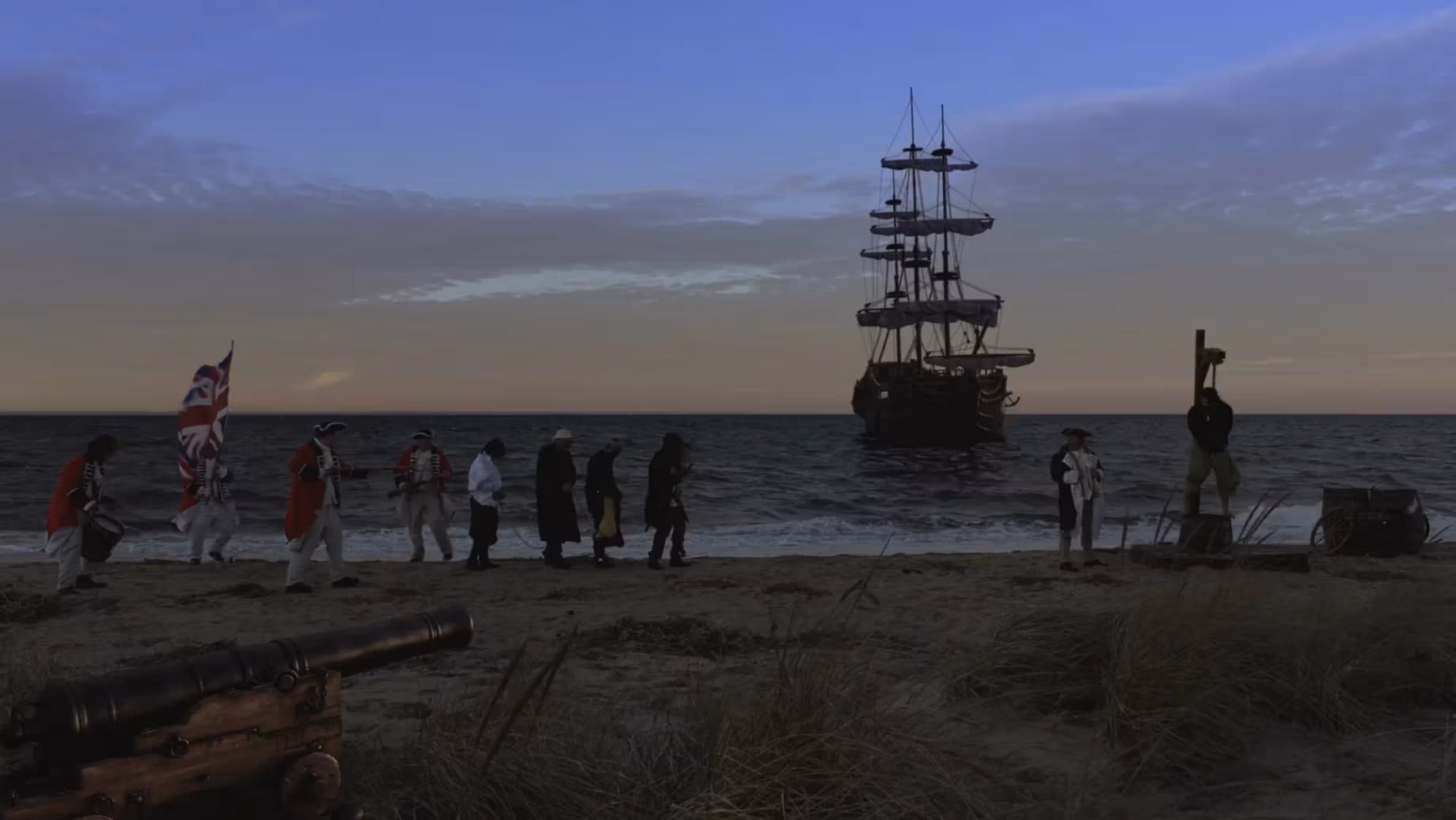 Dramatic sunset frame of a ship near shore; on the beach, a group of people walk, one carrying a union jack flag