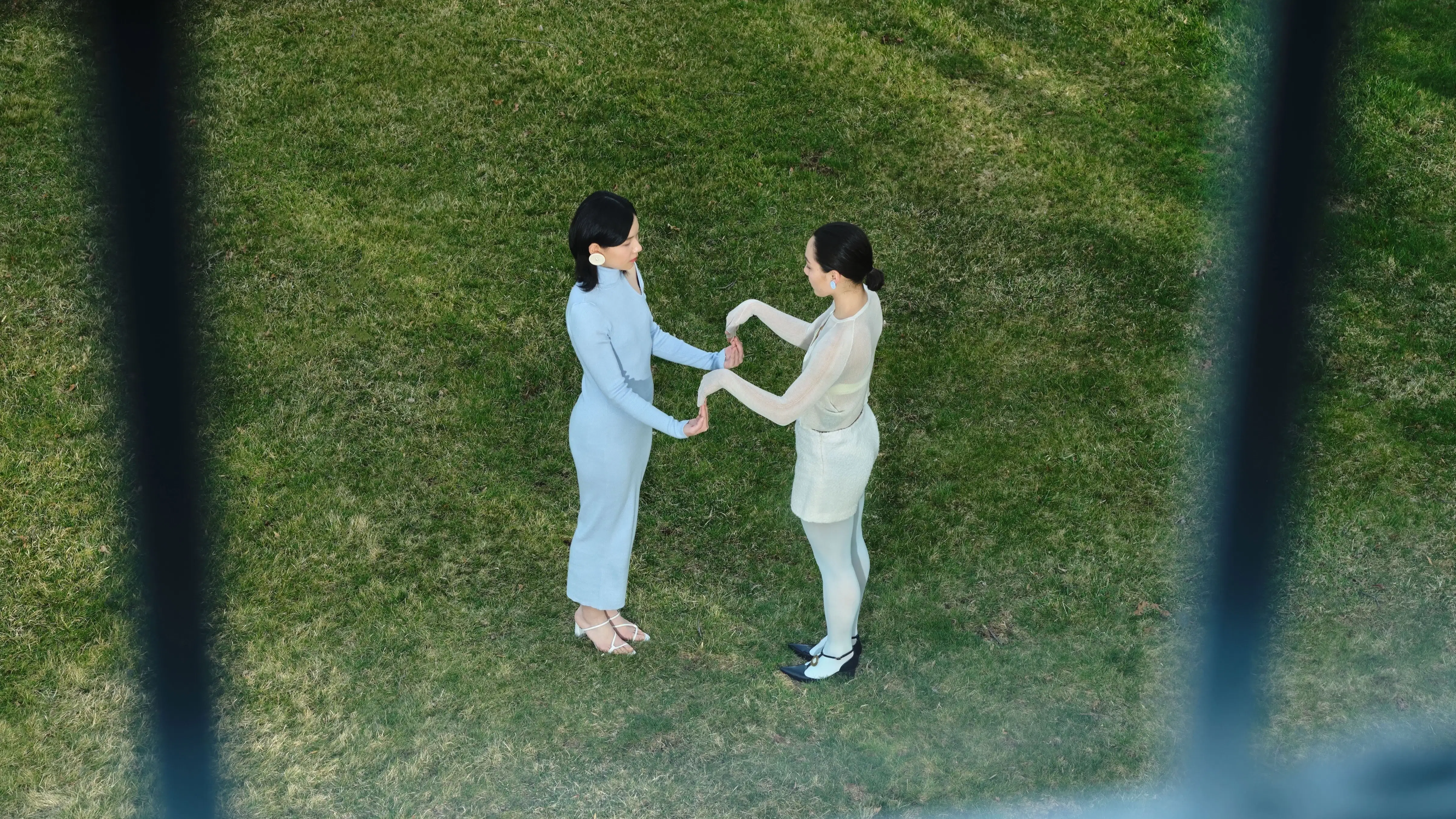 Birds eye view of Two people playing a hand clap game; the grass beneath their feet is very green.