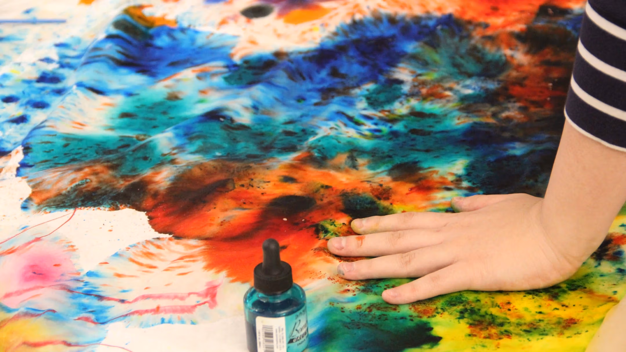 Photograph of a multicolored canvas on the floor with a hand atop supporting the weight of someone in the act of painting it
