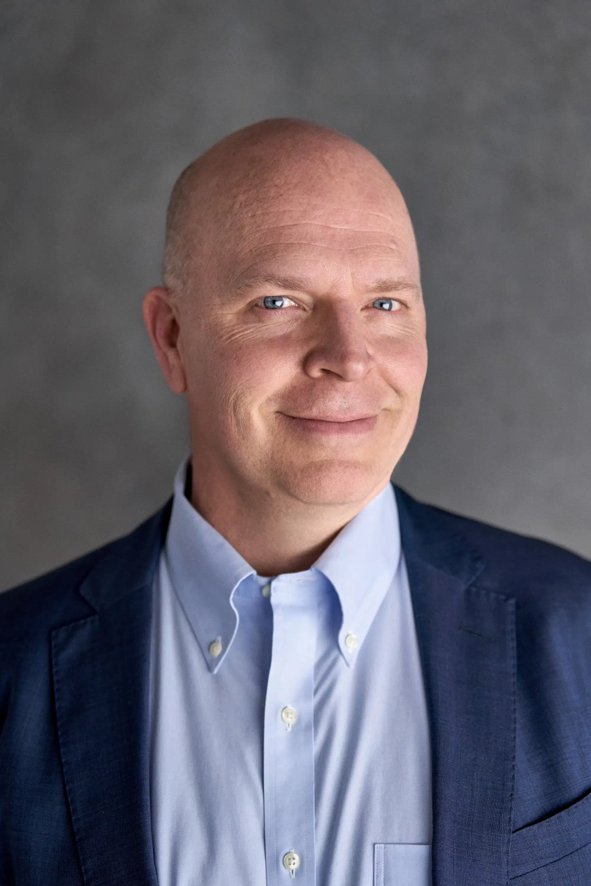  A middle-aged man with light skin and blue eyes, wearing a light blue button-down shirt and a navy blazer. He is smiling slightly and looking directly at the camera. The background is a neutral gray, creating a professional and approachable tone.