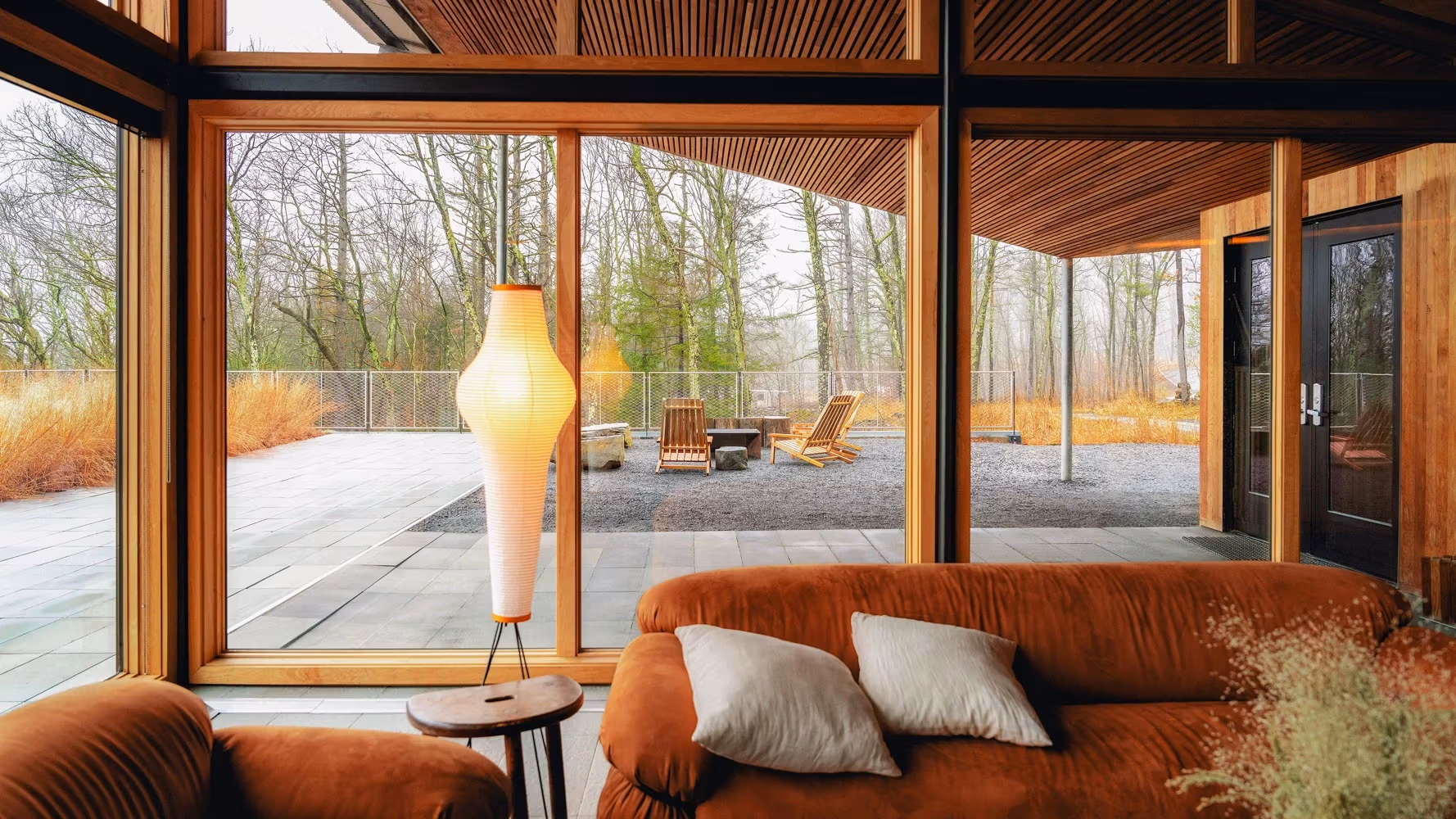 The interior of a wooden style resort reception area with a forest view. The overall color is warm orange. The foreground features two dark orange minimalist retro style matte fabric sofas, with a chocolate colored mini side table in between.